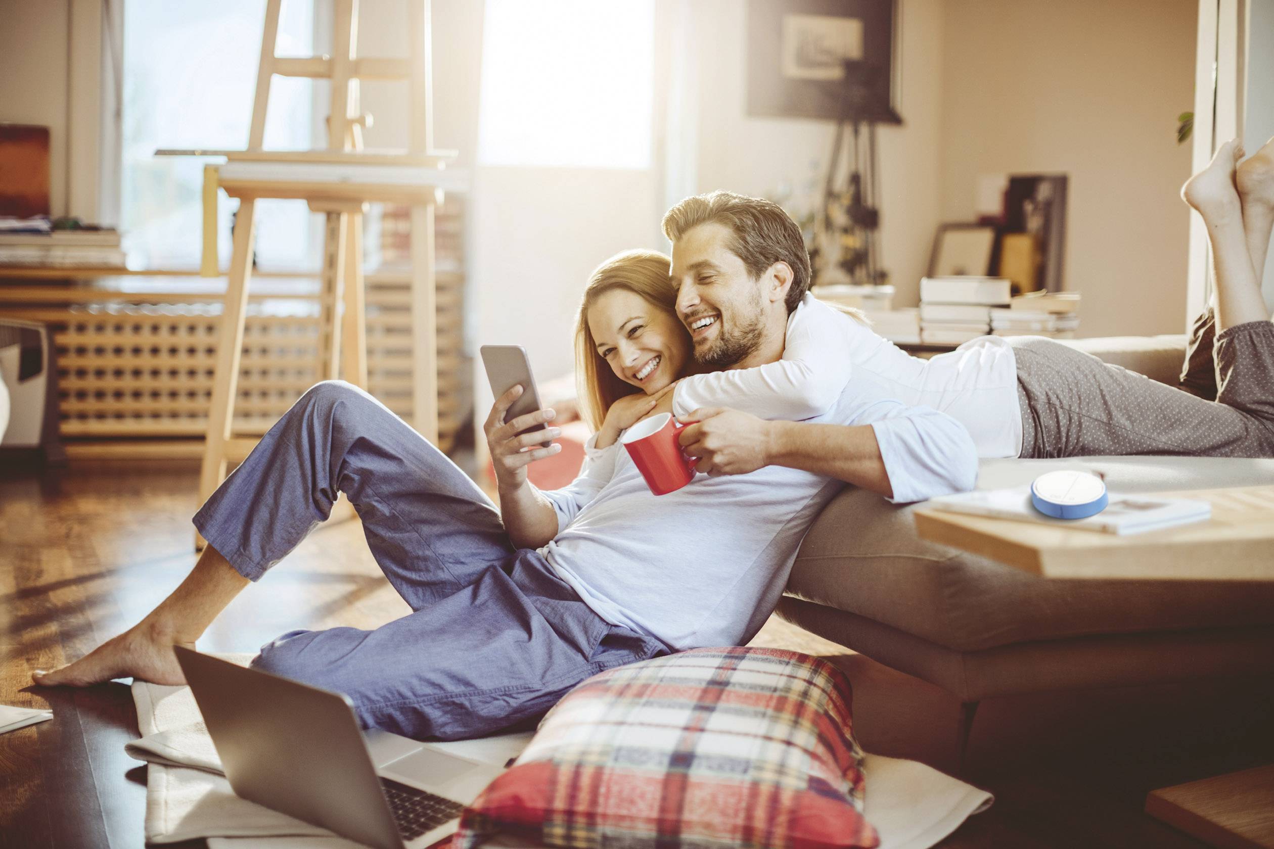 Para siedzi swobodnie w salonie, uśmiecha się i przygląda smartfonowi. Obok nich na podłodze leży otwarty laptop.
