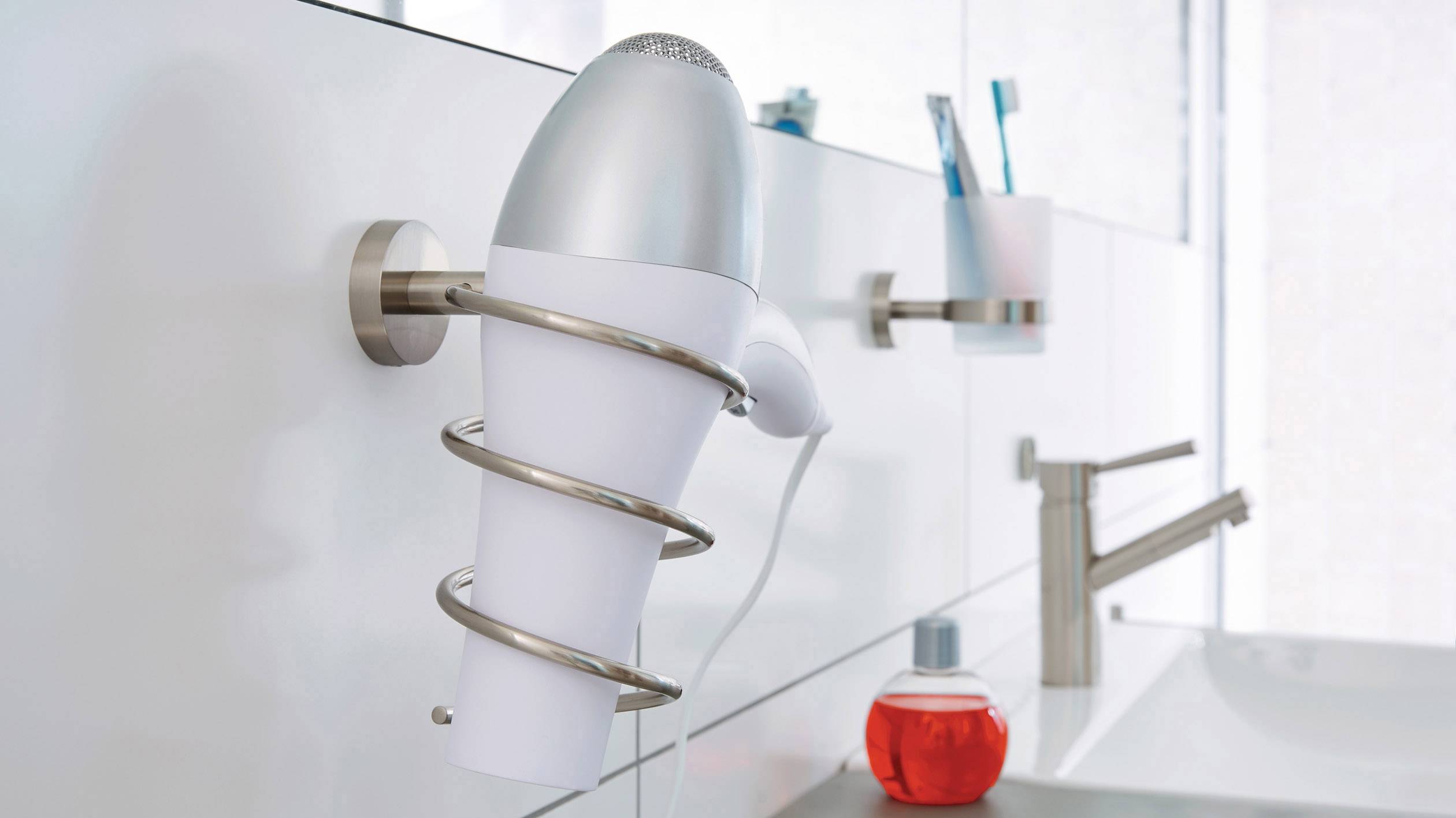 Suszarka do włosów wisi na ścianie w łazience. W tle widoczne są szczoteczki do zębów i czerwona butelka obok kranu.