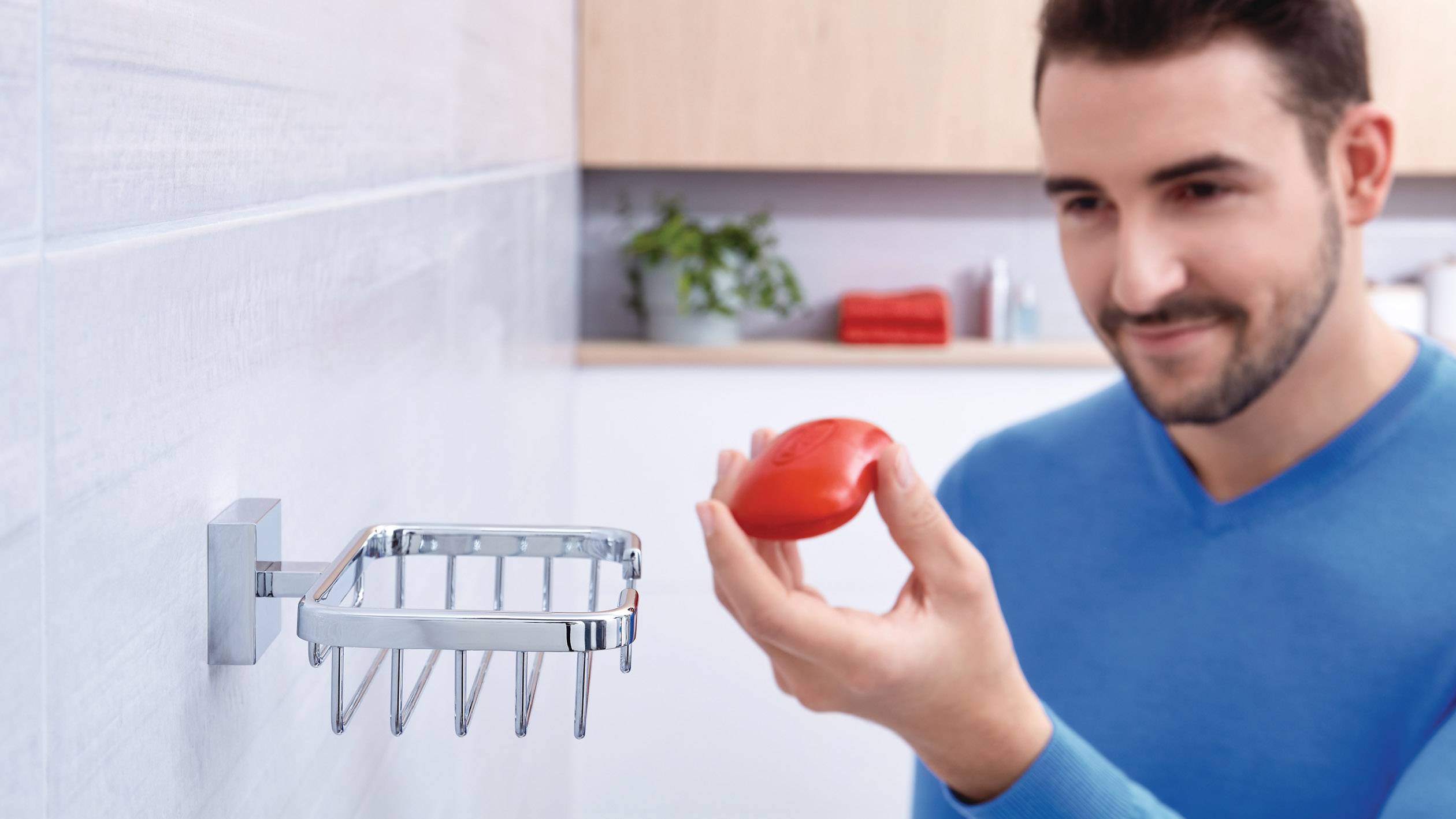 Mężczyzna w niebieskim swetrze stoi w łazience i trzyma w ręku kawałek czerwonego mydła. W tle widać różne akcesoria łazienkowe.