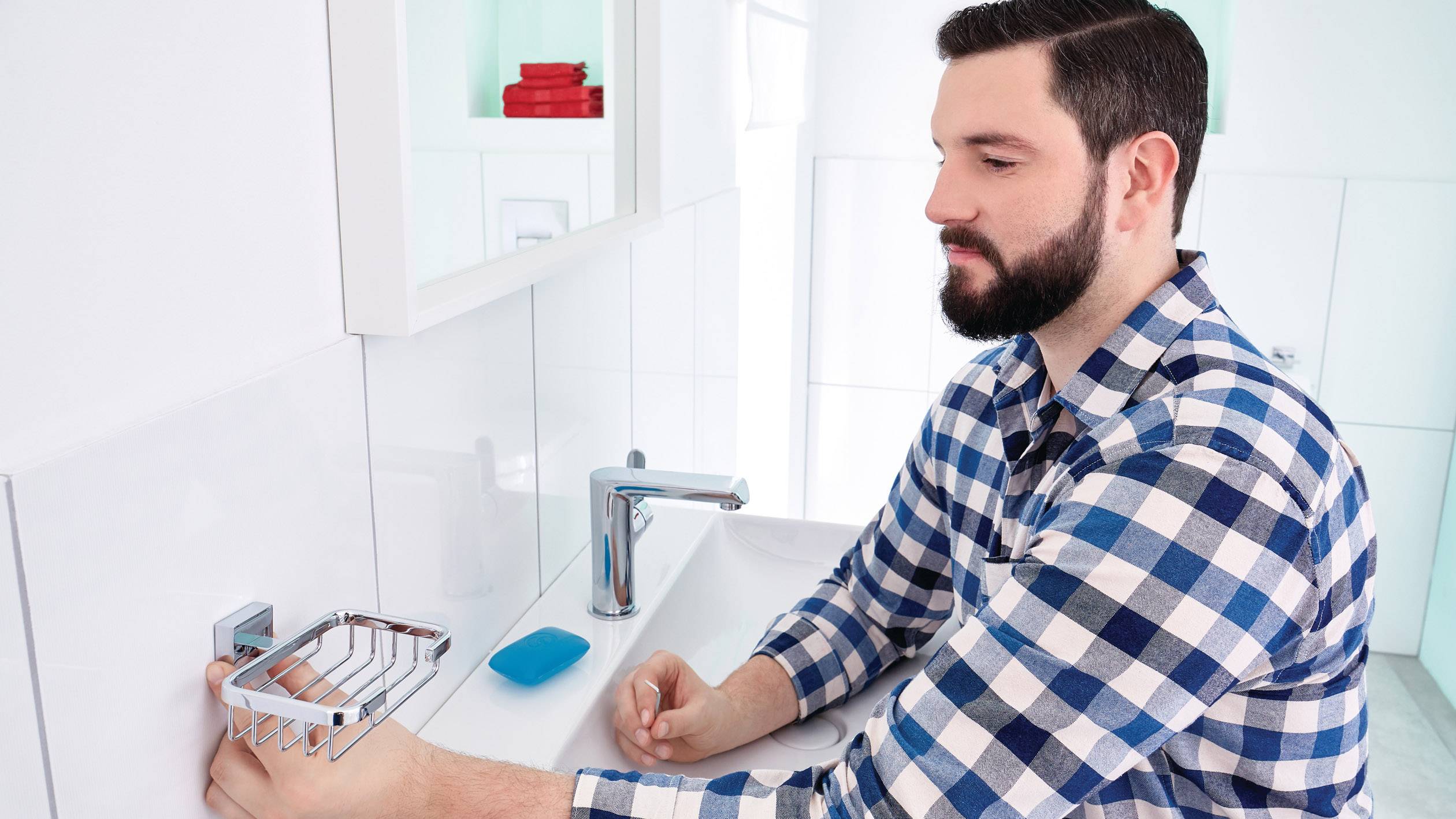 Mężczyzna z brodą w kraciastej koszuli myje ręce w łazience. Na tle widoczny jest dozownik mydła, niebieski ręcznik i czerwone ręczniki.