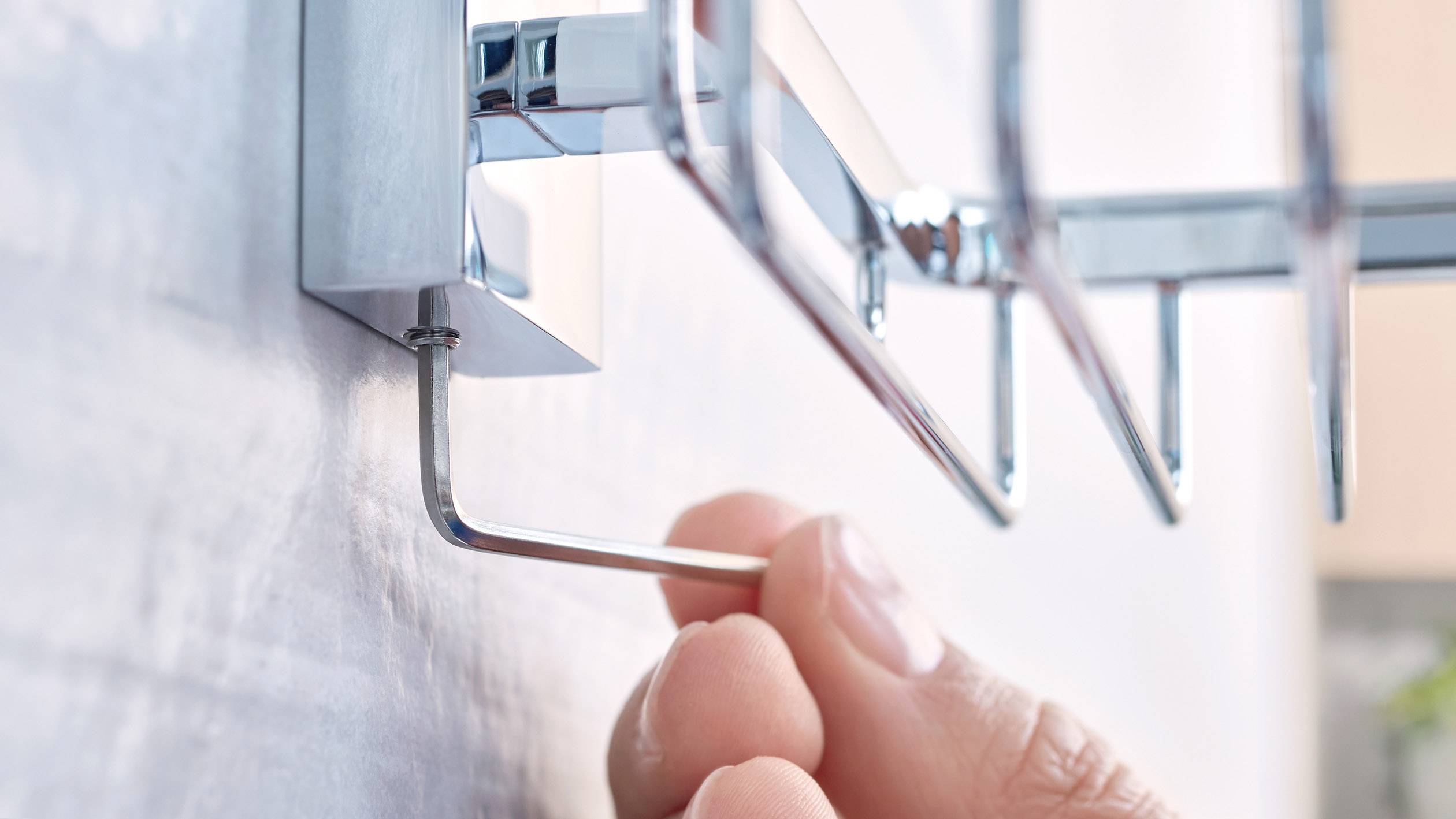 Ręka montuje chromowaną półkę ścienną za pomocą klucza imbusowego na ścianie.