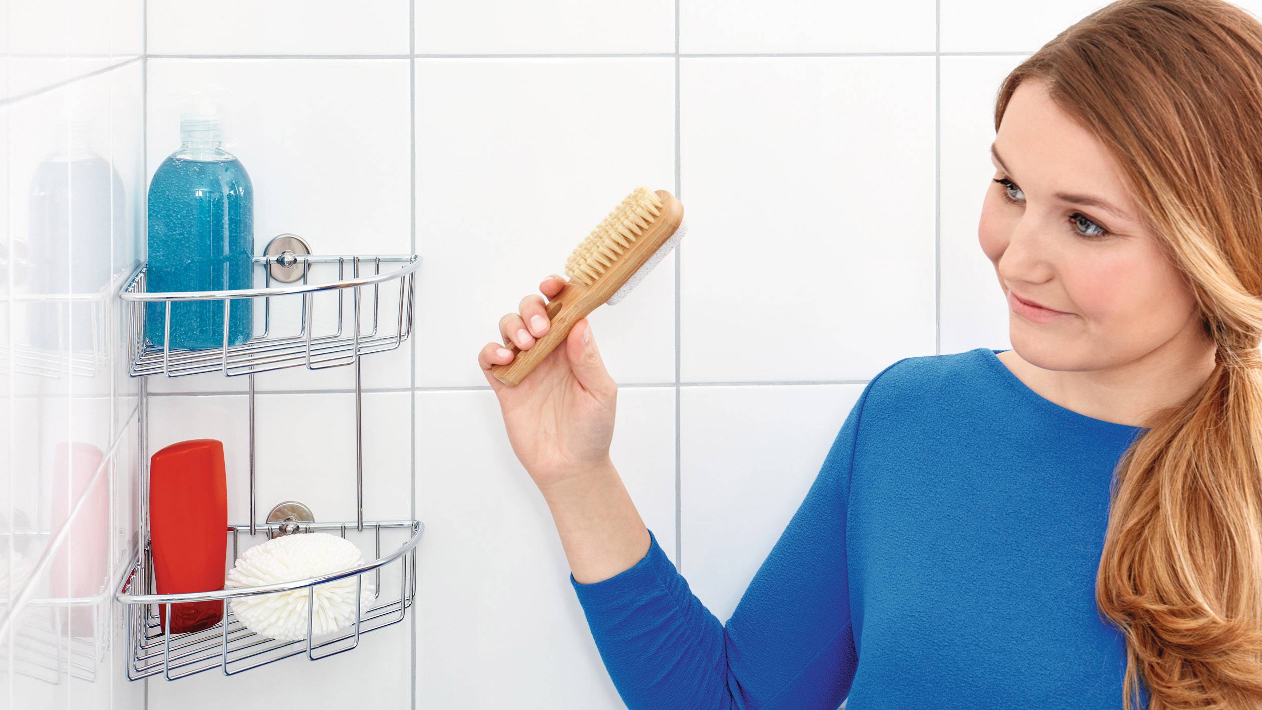 Kobieta w niebieskiej górze stoi w łazience i trzyma szczotkę. W tle na półce widoczne są produkty prysznicowe.