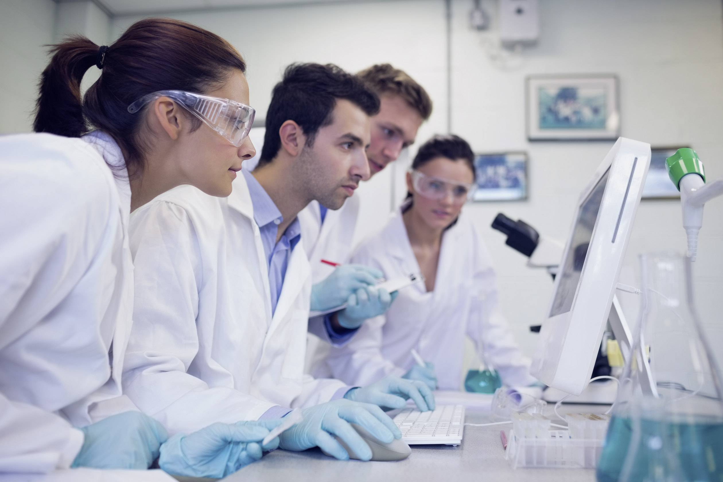 Czterech naukowców w białych fartuchach laboratoryjnych i okularach ochronnych pracuje przy komputerze w laboratorium, w tle widać probówki.