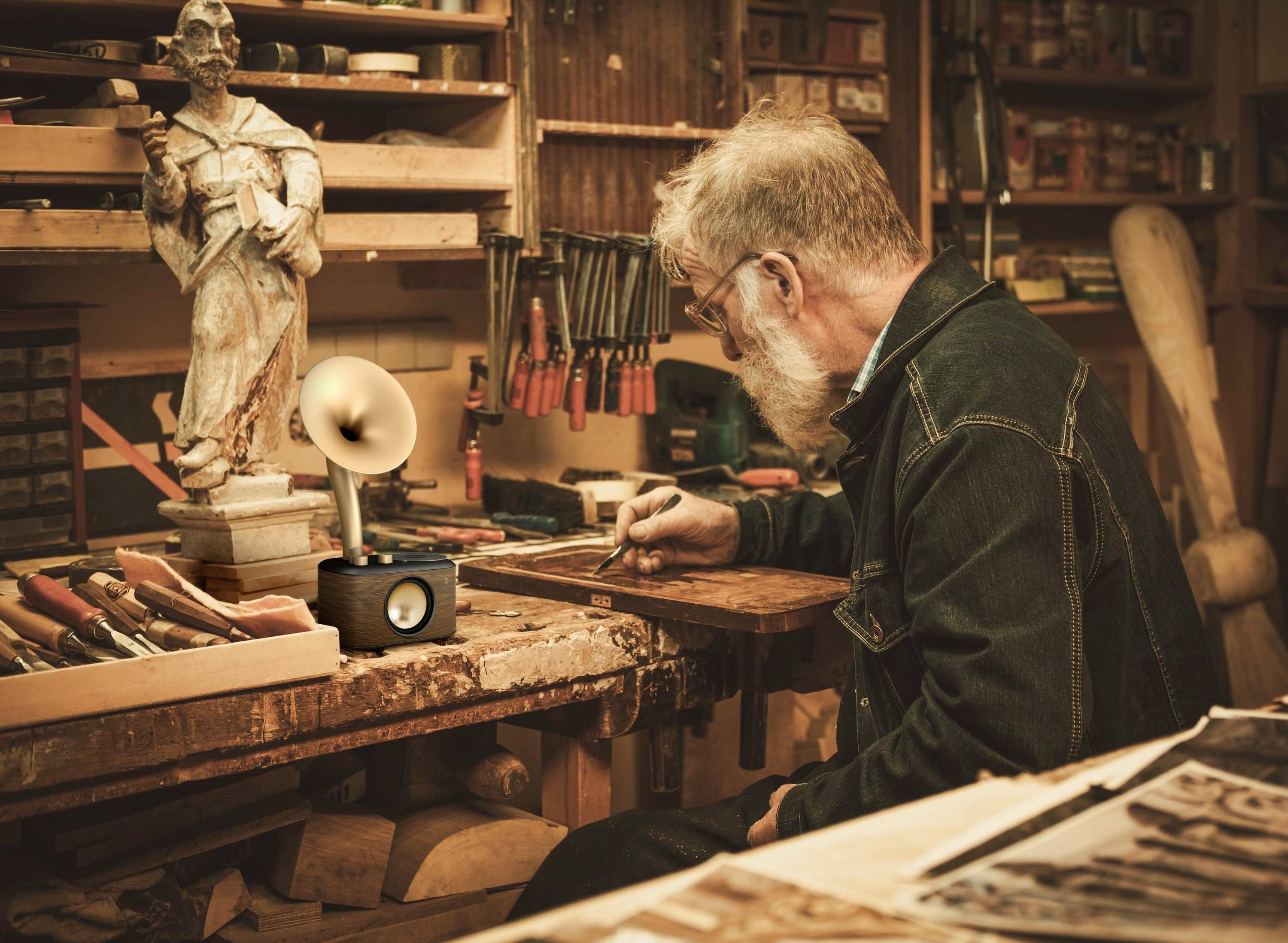 Mężczyzna z siwymi włosami i brodą pracuje przy stole w warsztacie, otoczony narzędziami i rzeźbą.