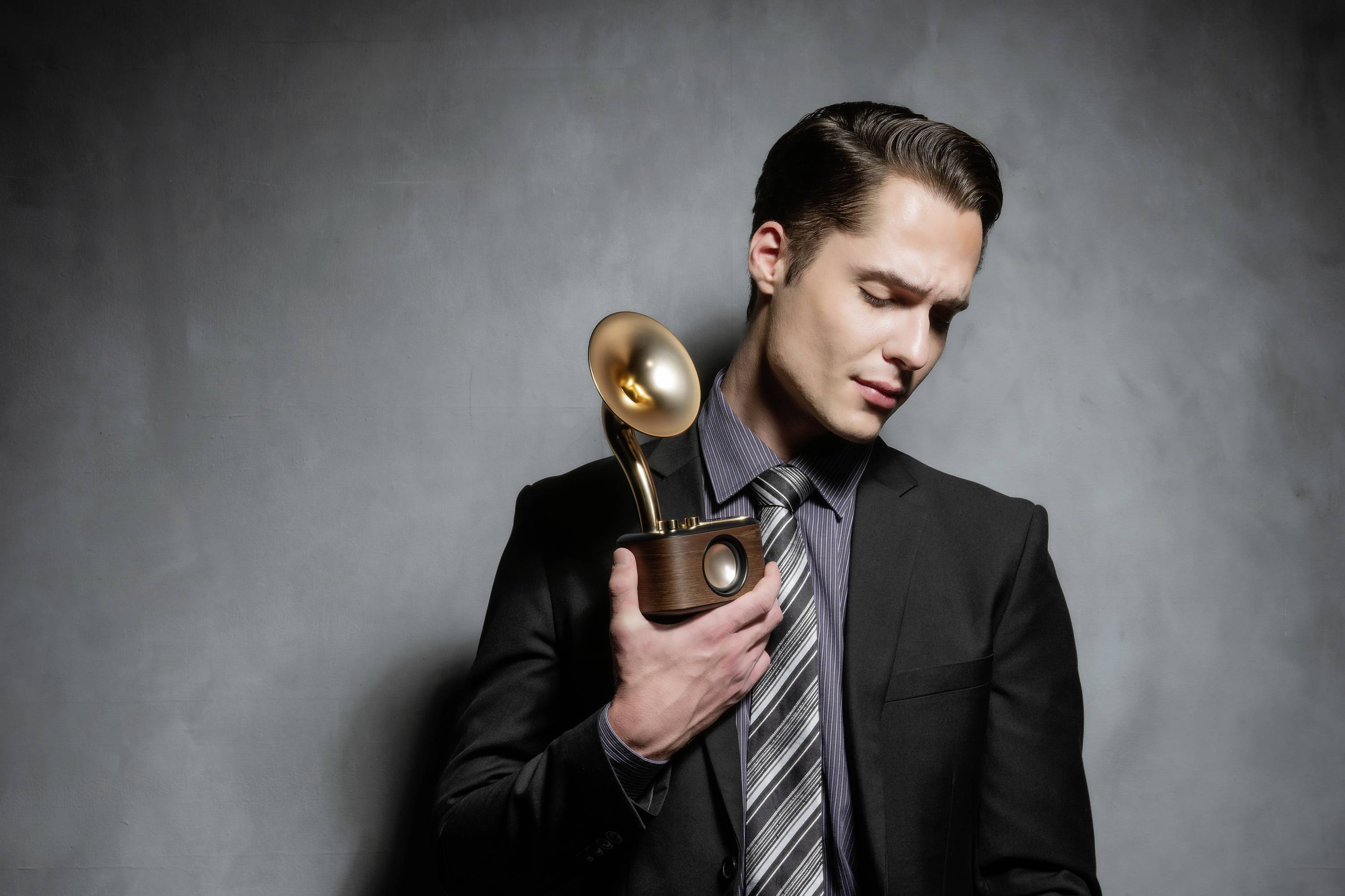 Mężczyzna w garniturze trzyma nagrodę gramofonu. Patrzy w dół i stoi na neutralnym tle.