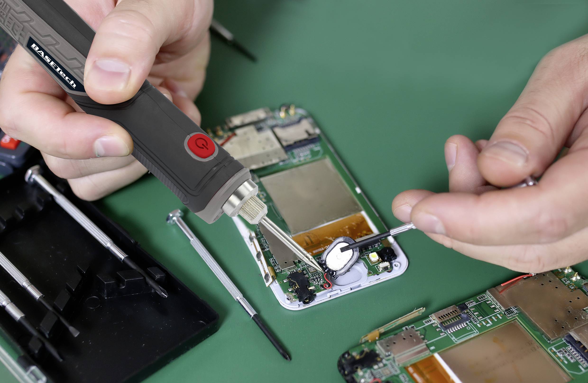 Dwie ręce naprawiają smartfon za pomocą lutownicy i narzędzi na stole. Widoczne są płytki elektroniczne i wkrętaki.
