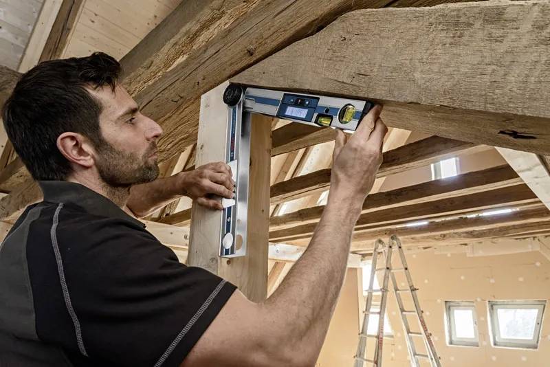 Osoba używa cyfrowego miernika kąta na drewnianej belce w częściowo wybudowanym pomieszczeniu, wskazując na precyzję w pracy stolarskiej.