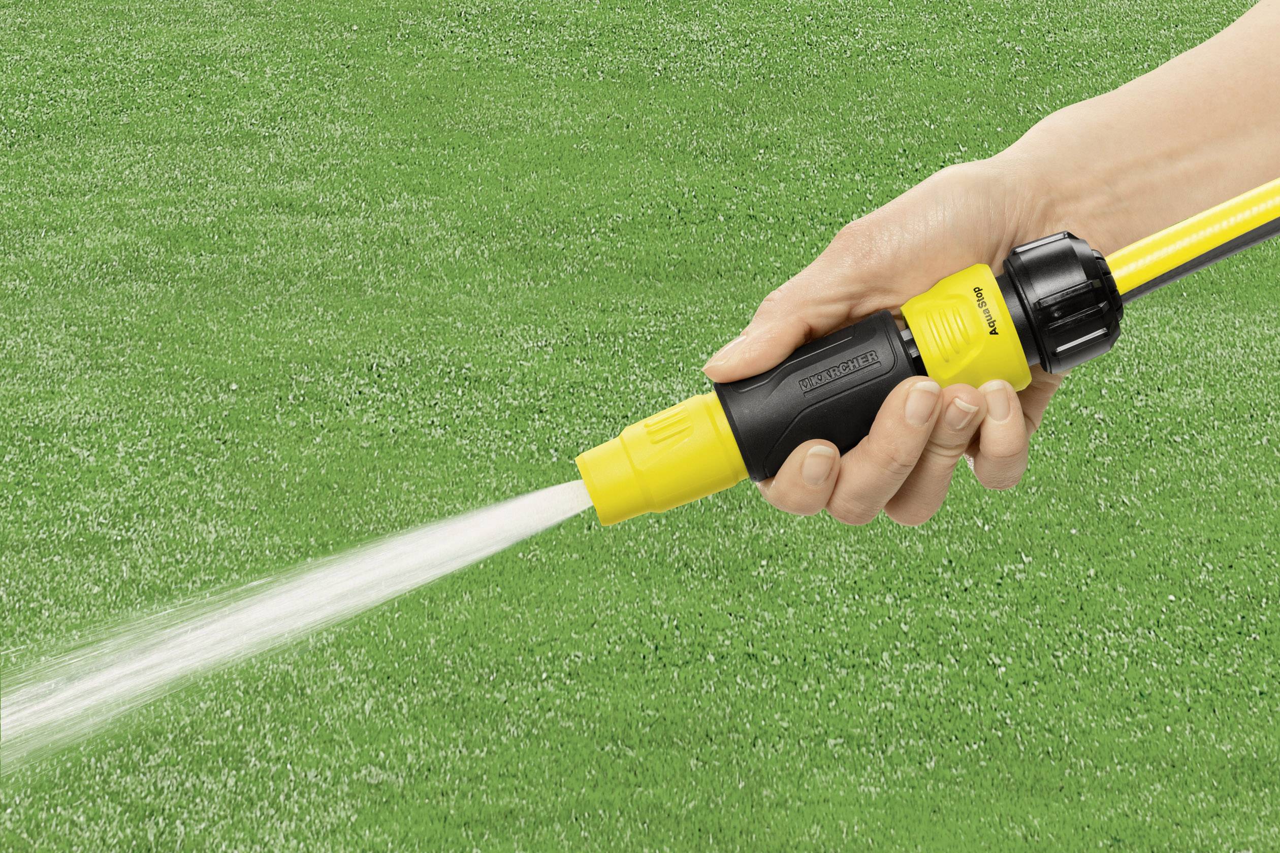 Dłoń trzyma wąż ogrodowy z dyszą, z której woda jest rozpryskiwana na zielony trawnik.