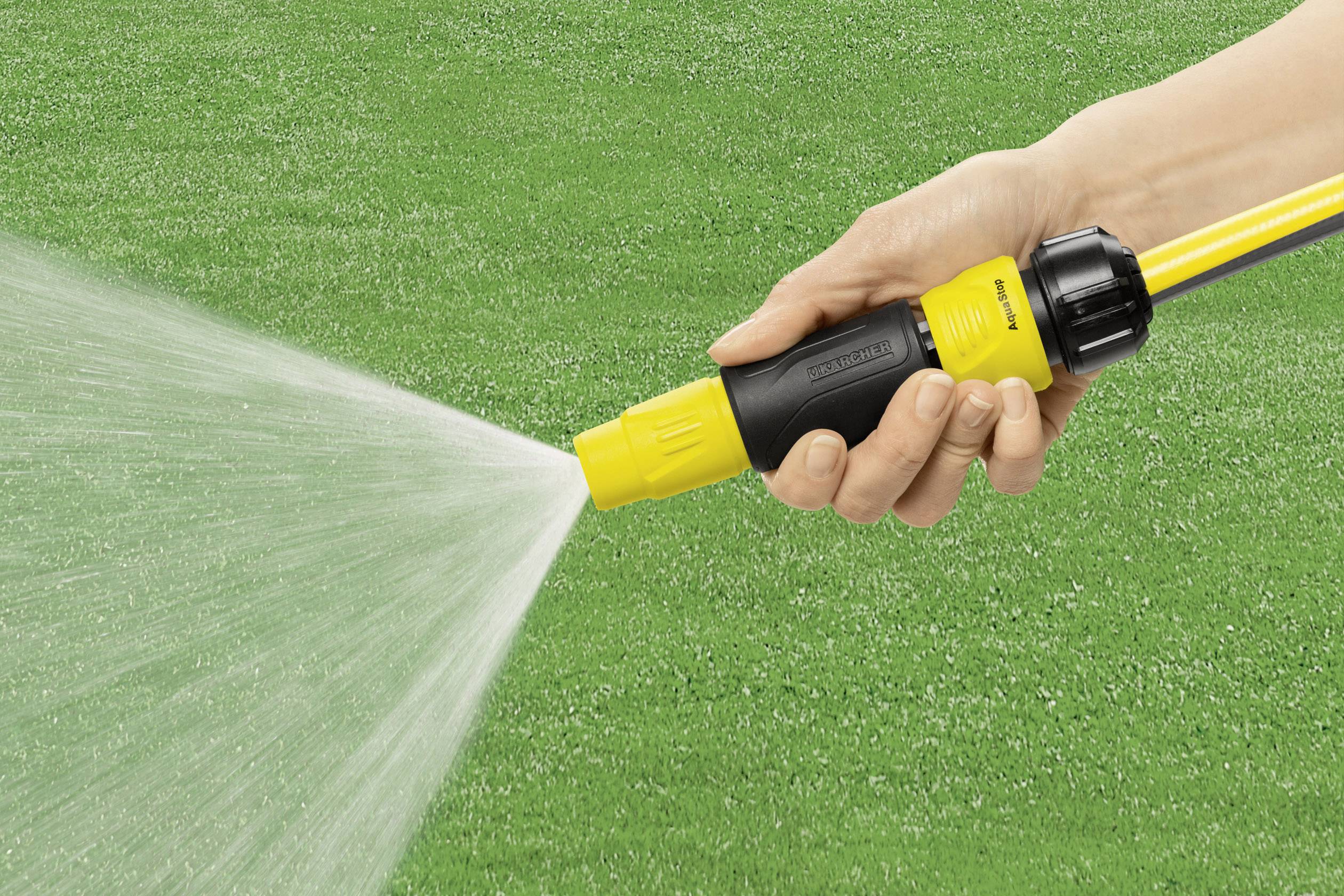 Ręka trzyma dyszę węża ogrodowego, która rozpyla wodę na zielony trawnik.