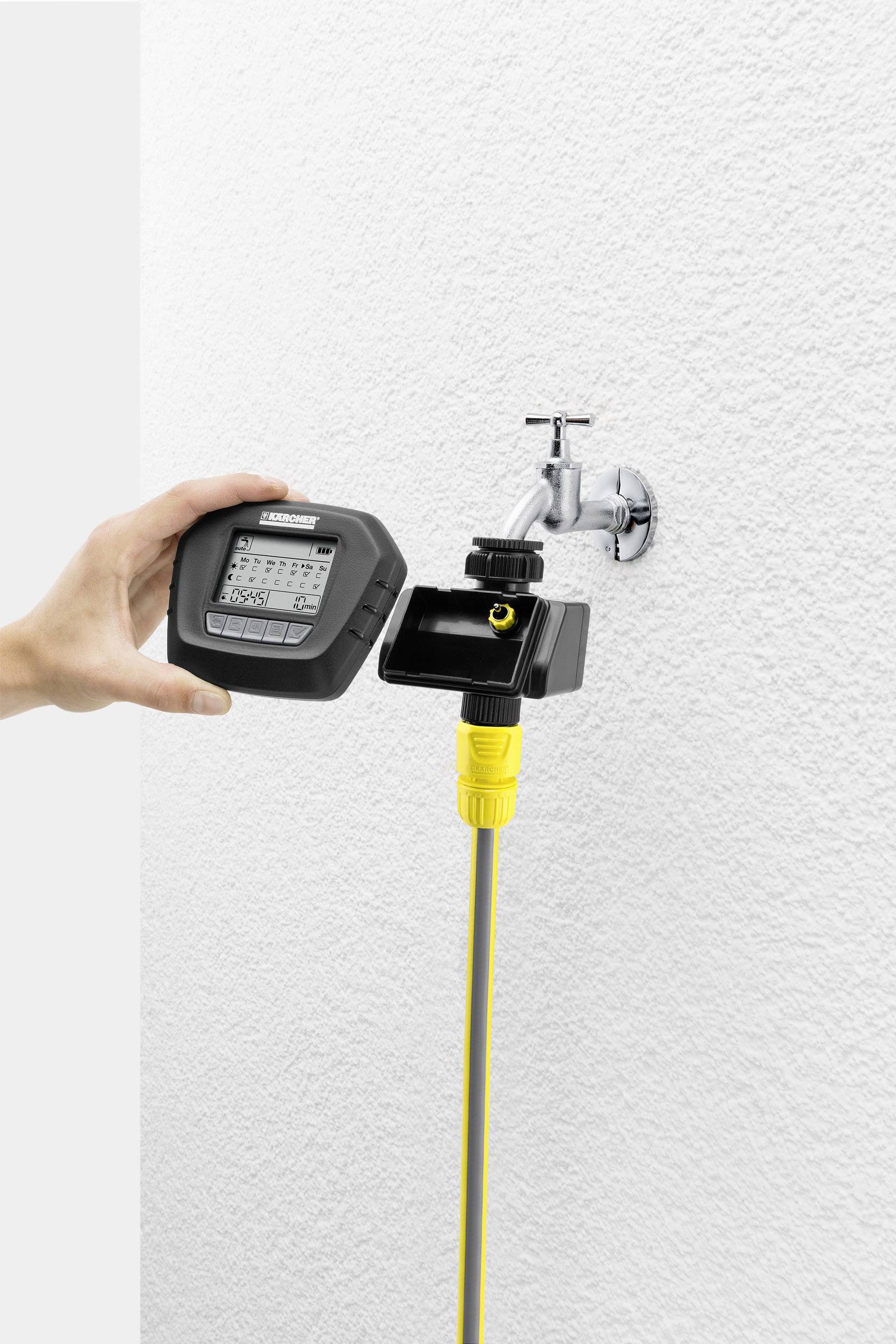 Ręka trzyma urządzenie do sterowania nawadnianiem obok kranu z podłączonym żółtym wężem przy białej ścianie.