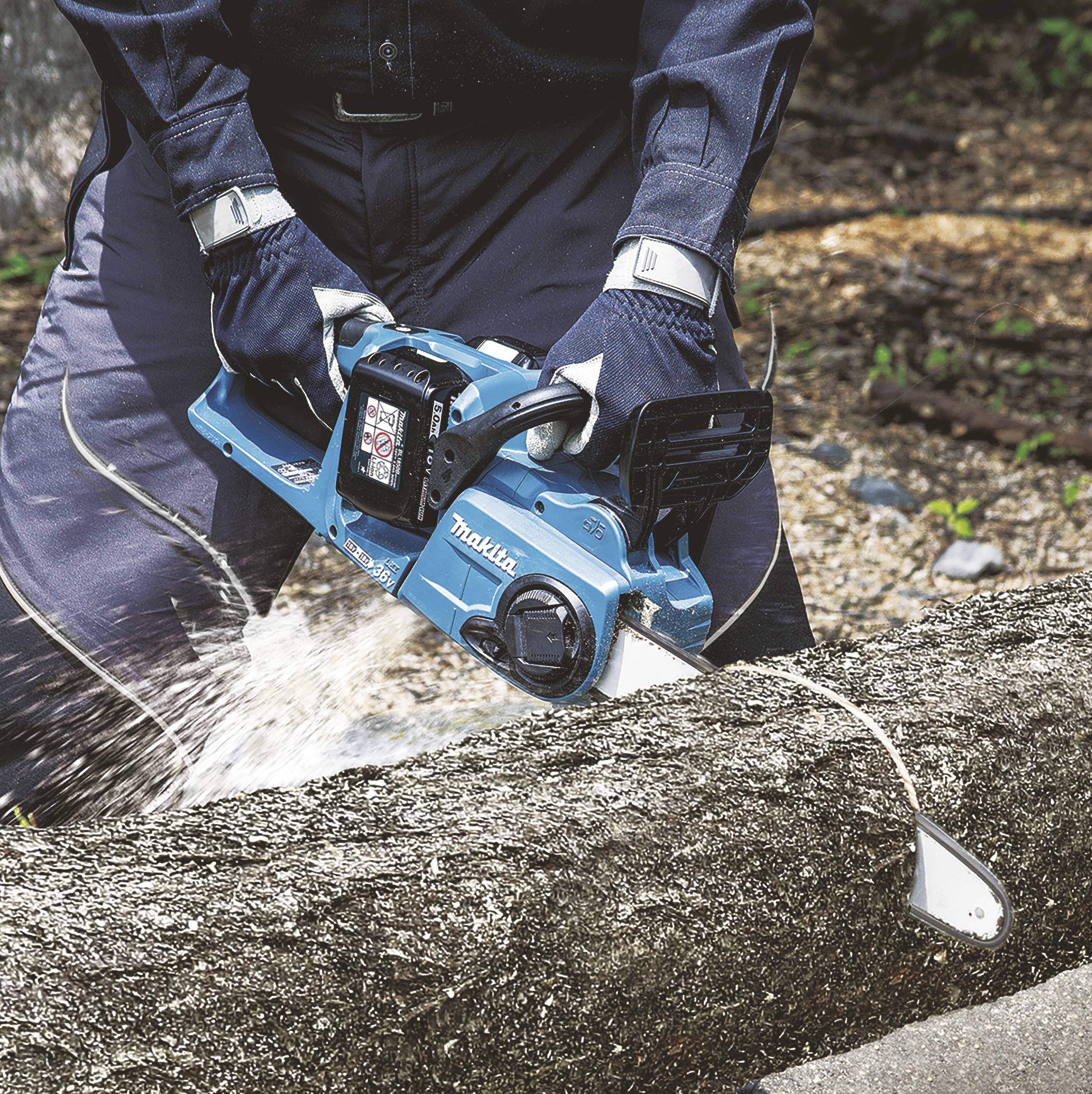 Osoba piłuje niebieską elektryczną piłą łańcuchową pień drzewa. Wióry odlatują na boki, otoczenie jest zalesione.