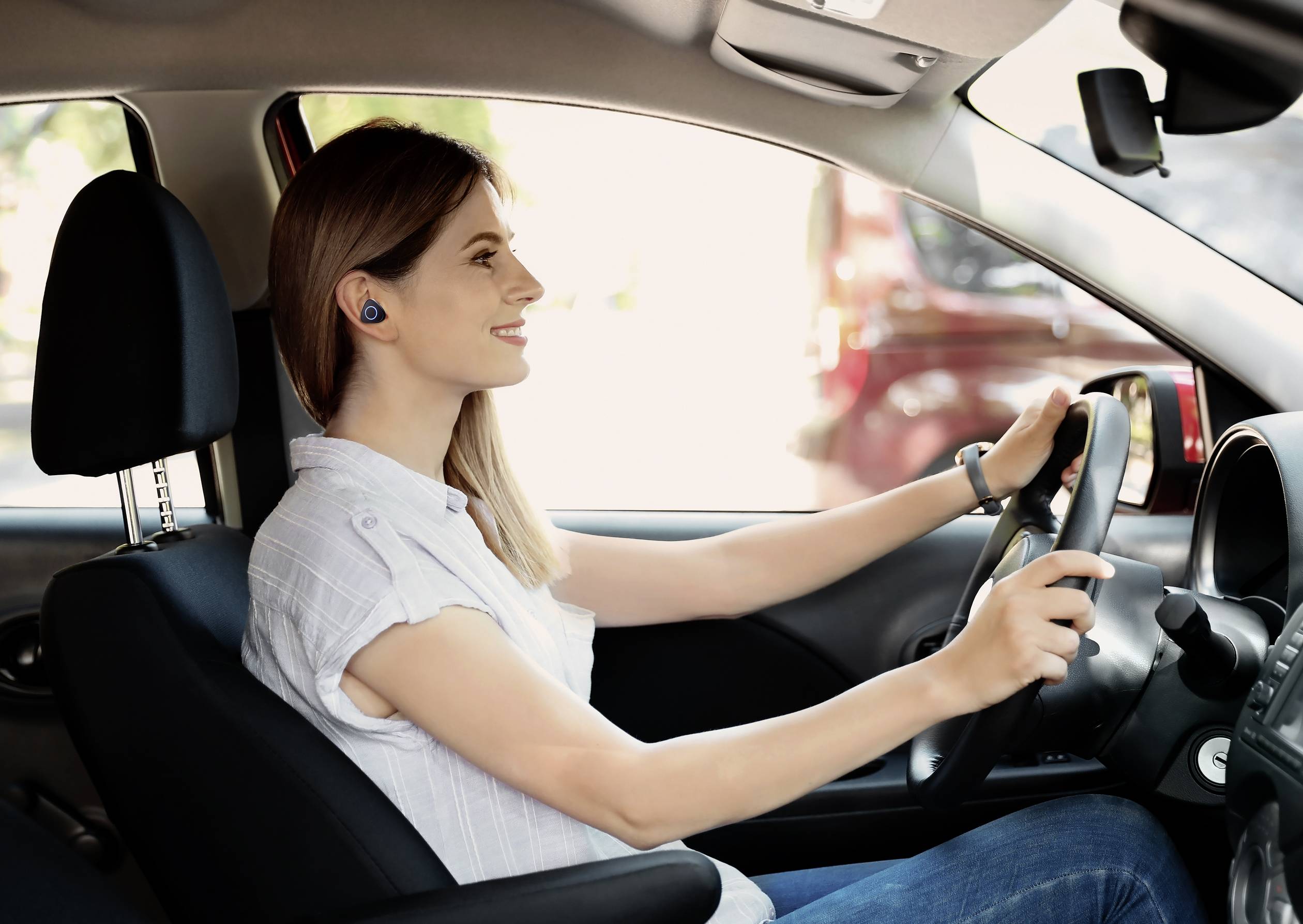 Uśmiechnięta osoba prowadzi samochód i nosi bezprzewodowe słuchawki douszne. Siedzi za kierownicą i jest skoncentrowana na drodze.