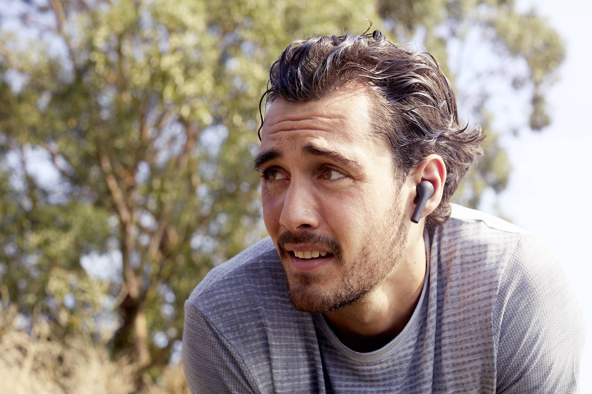 Młody mężczyzna z brodą w sportowej koszulce i bezprzewodowych słuchawkach, który zdaje się być na zewnątrz, z drzewami w tle.
