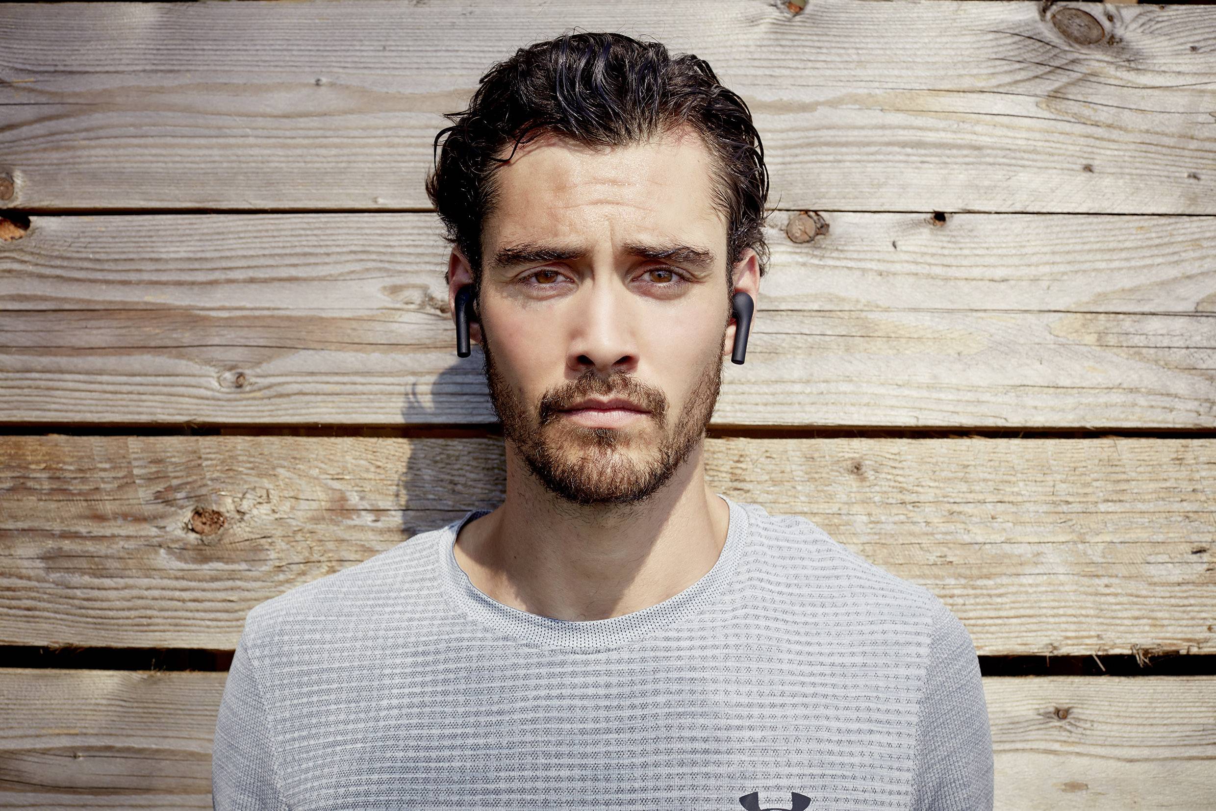 Młody mężczyzna z brodą nosi bezprzewodowe słuchawki douszne i poważnym wzrokiem patrzy w obiektyw. Tło stanowi drewniana ściana.