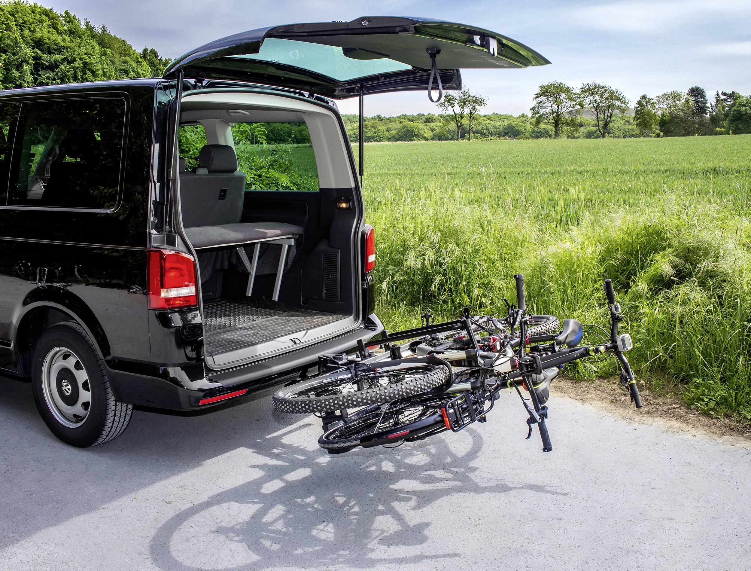 Otwarty bagażnik vana z wysuwanym bagażnikiem rowerowym, na którym zamontowane są dwa rowery, w tle zielone pola.