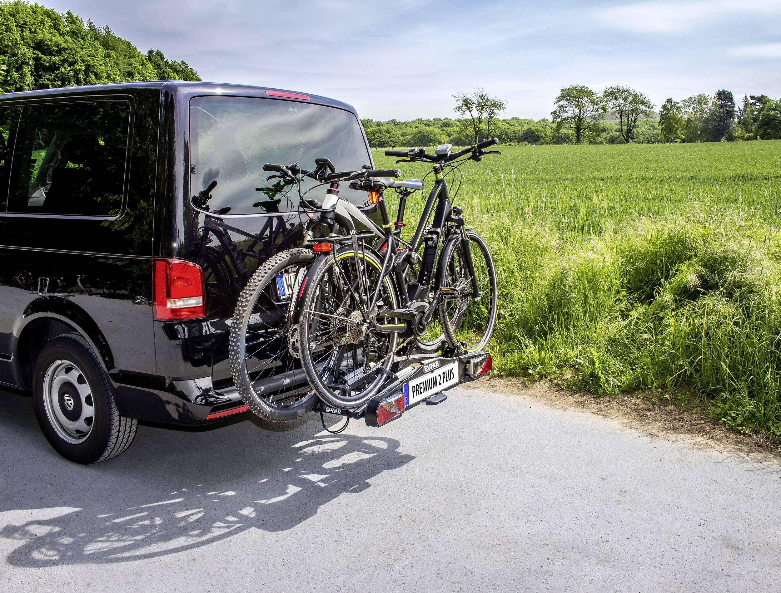 Czarny van z trzema rowerami na bagażniku tylnym, zaparkowany na drodze krajowej obok zielonego pola i drzew w tle.
