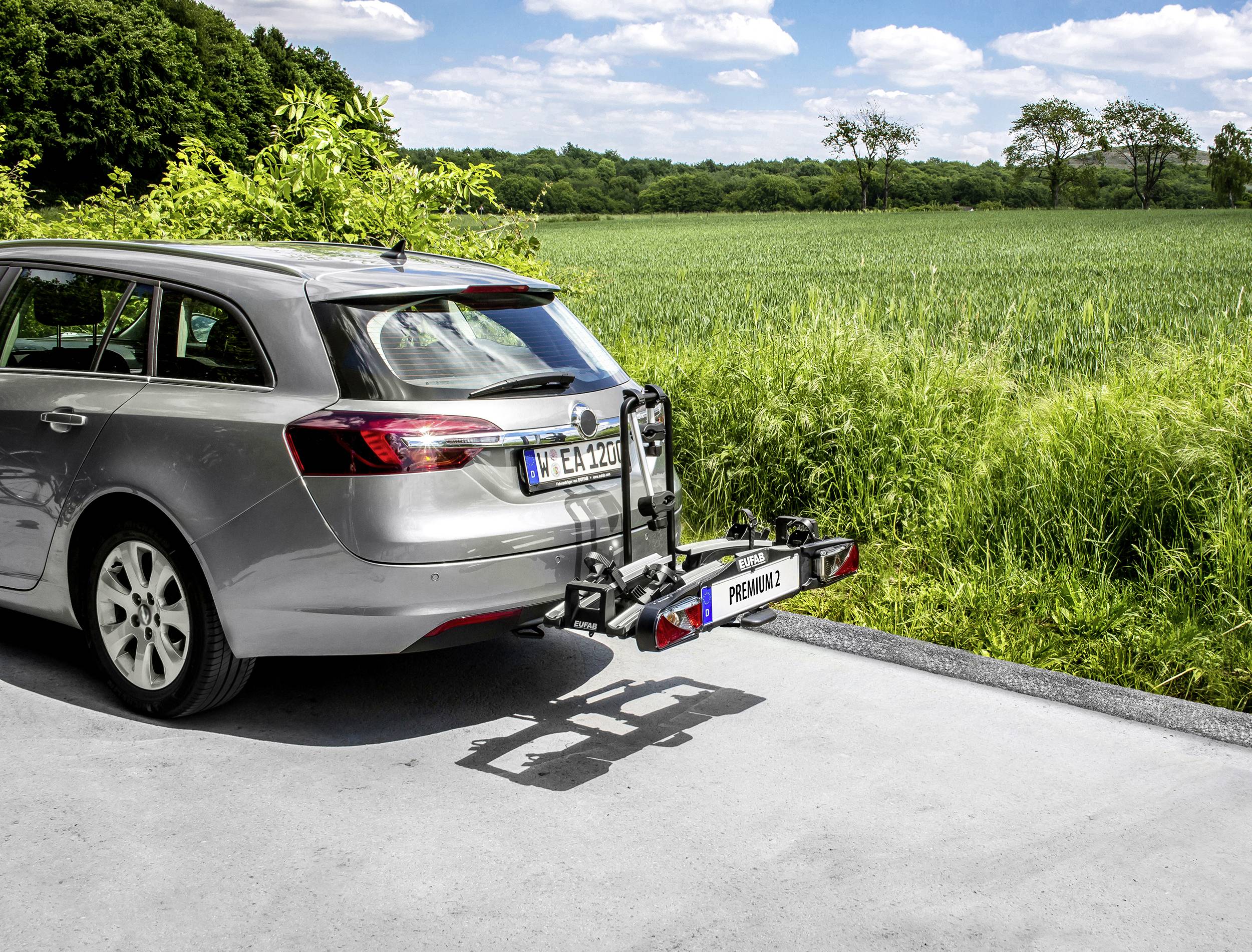 Srebrny samochód stoi na drodze obok zielonego pola. Z tyłu zamontowany jest bagażnik rowerowy, który jest pusty.