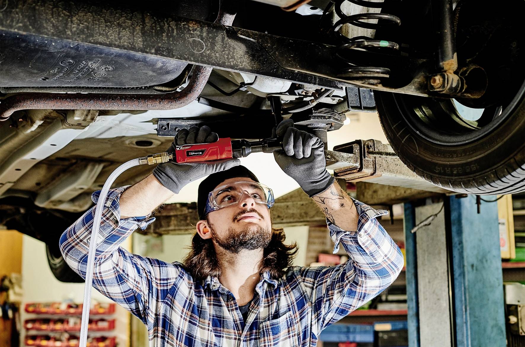 Mechanik naprawia podwozie samochodu w warsztacie, nosi okulary ochronne i używa narzędzia pneumatycznego.