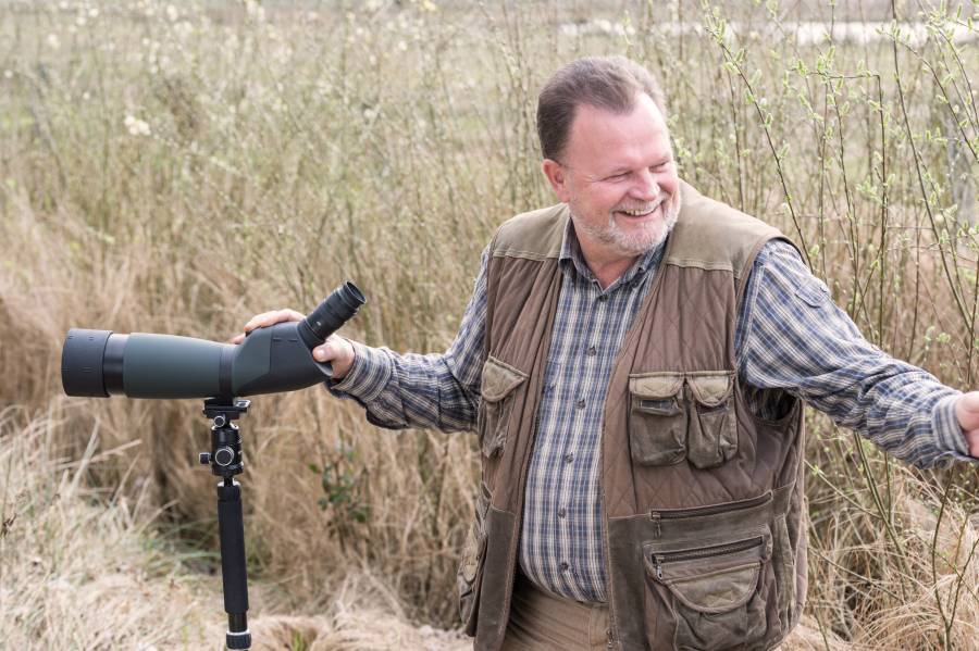 Mężczyzna na wolnym powietrzu uśmiecha się, trzymając teleskop na statywie, otoczony wysoką trawą.
