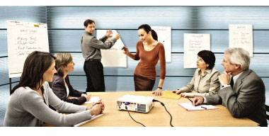 Sześć osób dyskutuje w sali konferencyjnej przed tablicą. Dwie osoby stoją i piszą, podczas gdy cztery siedzą przy stole. Na stole stoi projektor.