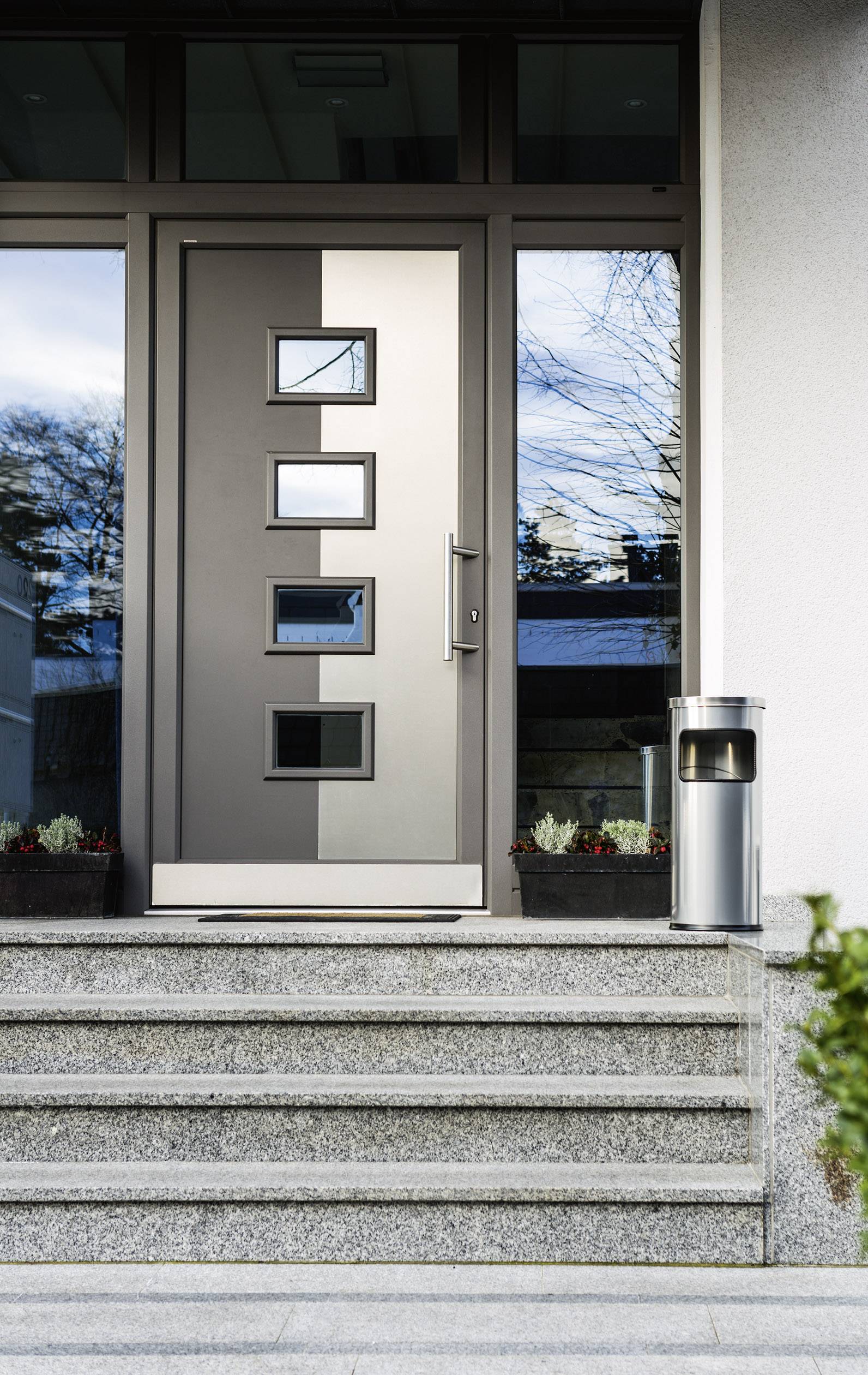 Nowoczesne drzwi wejściowe z czterema kwadratowymi oknami w szarej ramie. Po lewej i prawej stronie na schodach stoją doniczki z kwiatami.