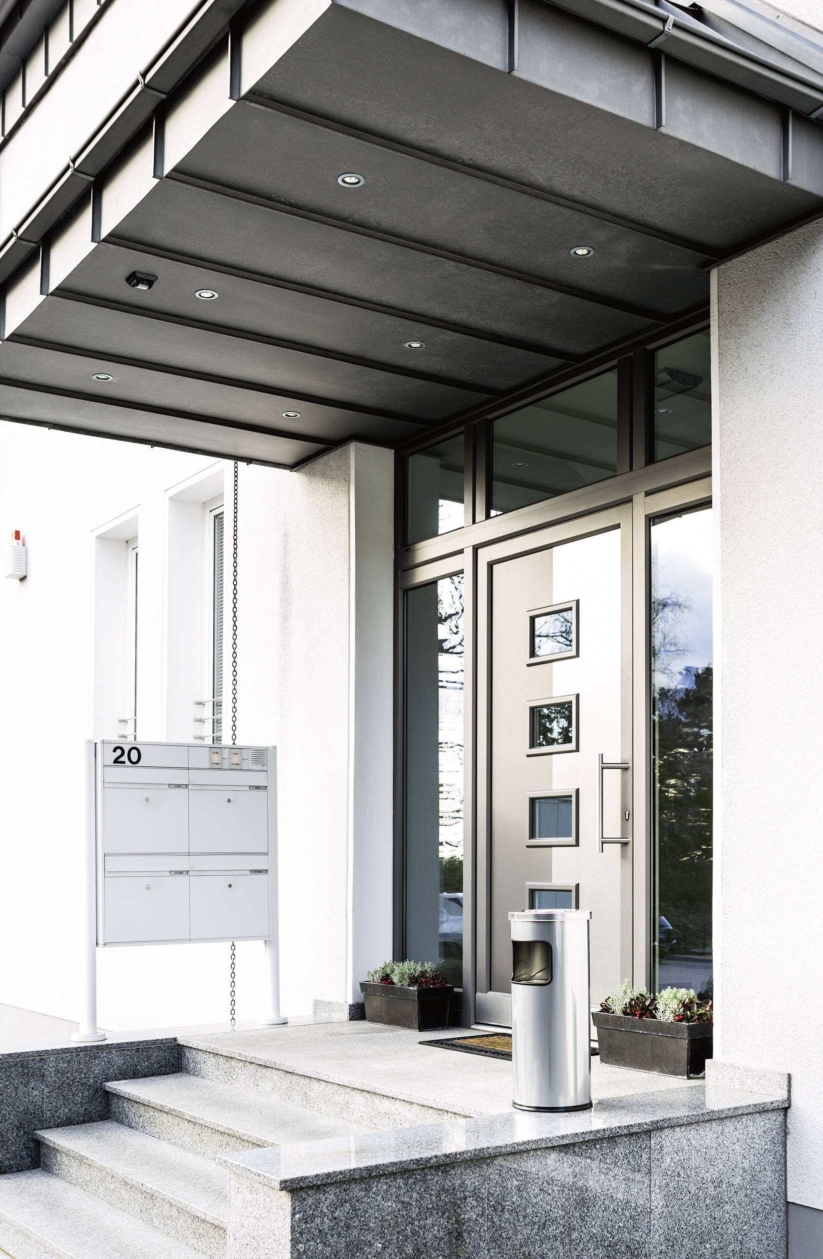 Nowoczesne wejście do domu z srebrną furtką, skrzynką na listy z numerem '20' i pojemnikiem na śmieci. Gładkie betonowe stopnie i zadbane rośliny.