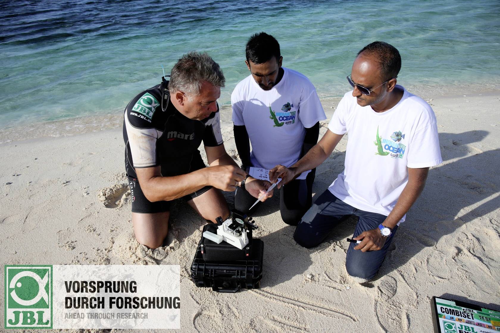 Trzech mężczyzn na plaży bada próbki. Jeden jest ubrany w skafander neoprenowy, pozostali dwaj w koszulkach. Na piasku leży walizka z wyposażeniem.