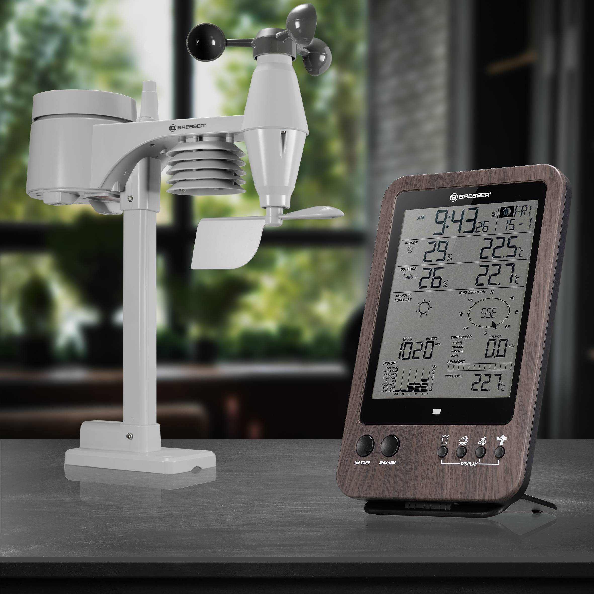 Cyfrowy wyświetlacz pogodowy z wskazaniem temperatury i wilgotności stoi na stole, obok urządzenia pomiarowego. Tło rozmyte.