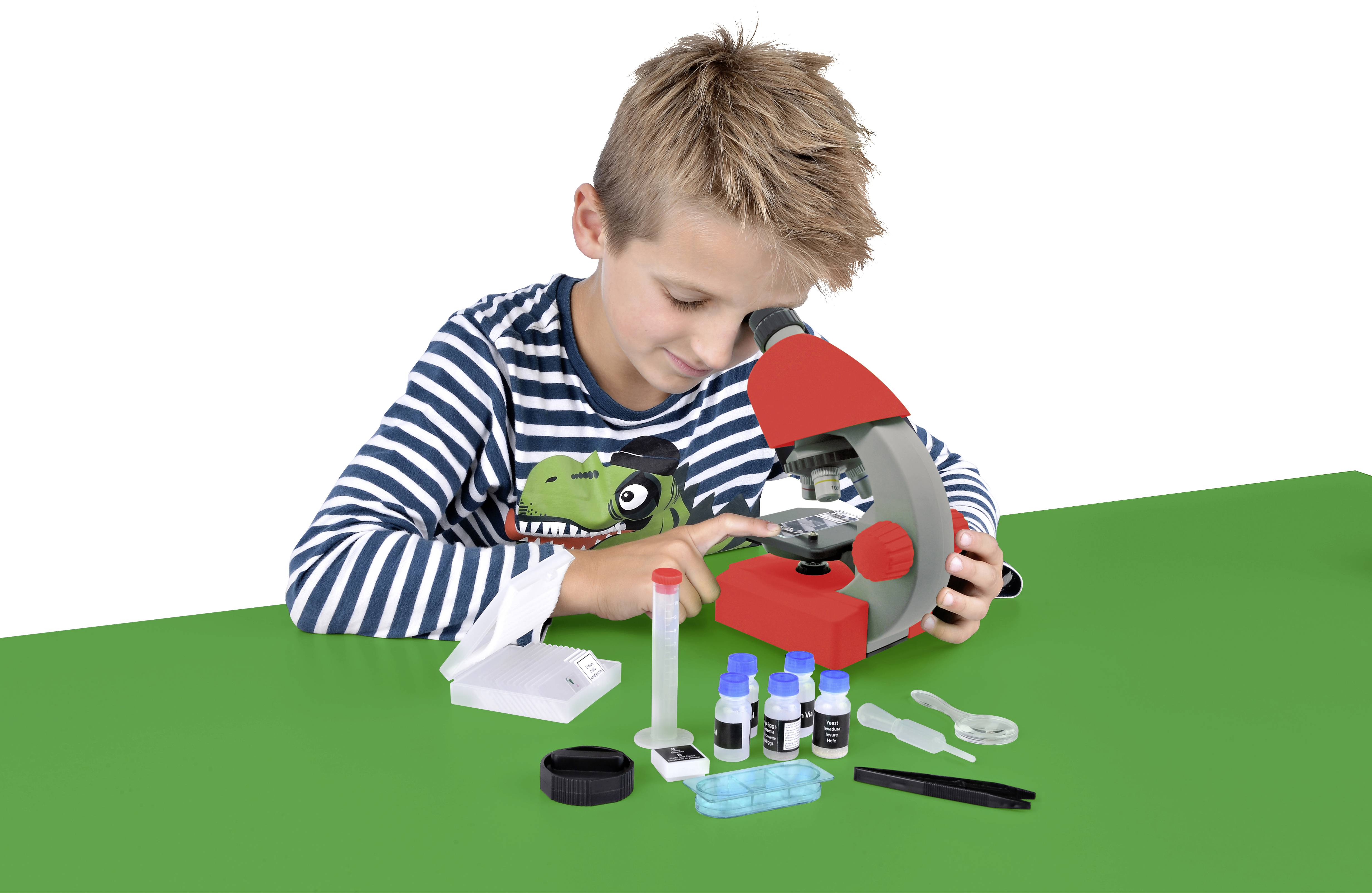 Chłopiec patrzy przez mikroskop. Na stole leżą różne przybory laboratoryjne, w tym fiolki i narzędzia.