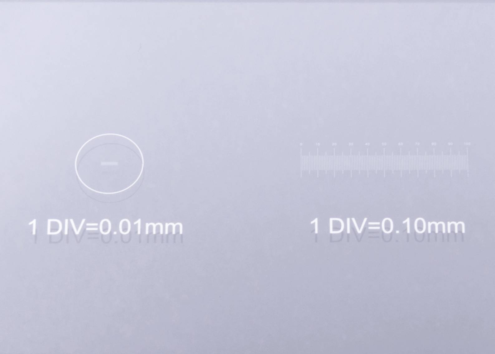 Szklany przyrząd pomiarowy z dwoma skalami: po lewej stronie okrągła skala z oznaczeniem '1 DIV=0,01mm', po prawej liniowa skala z oznaczeniem '1 DIV=0,10mm'.