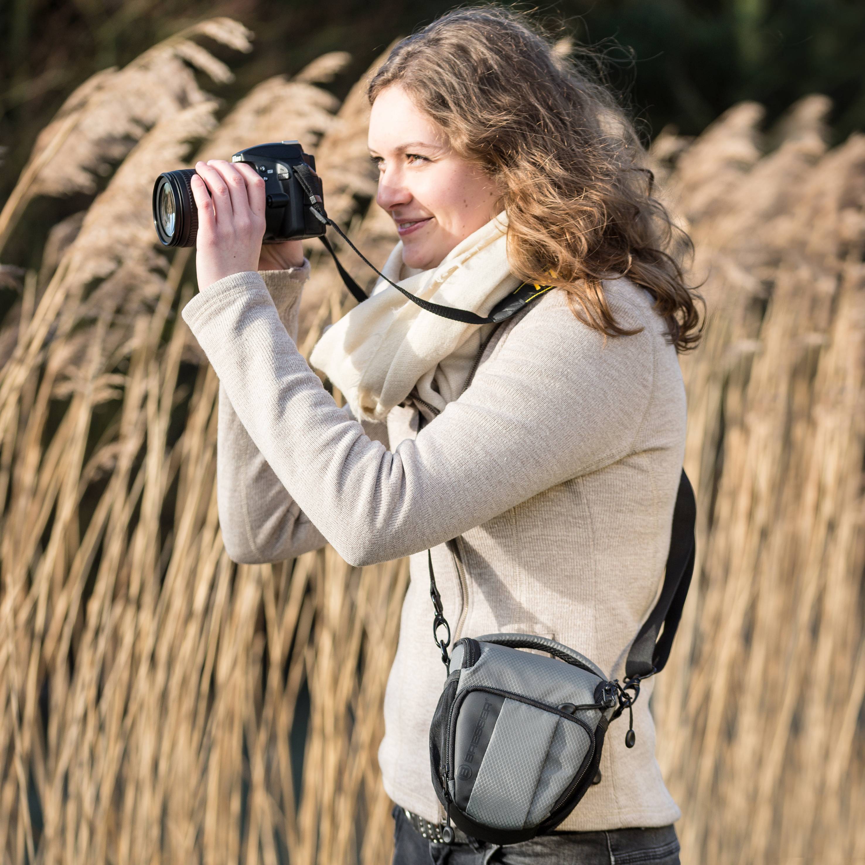 Kobieta robi zdjęcia w plenerze aparatem fotograficznym. W tle widać trzcinę kołyszącą się na wietrze.