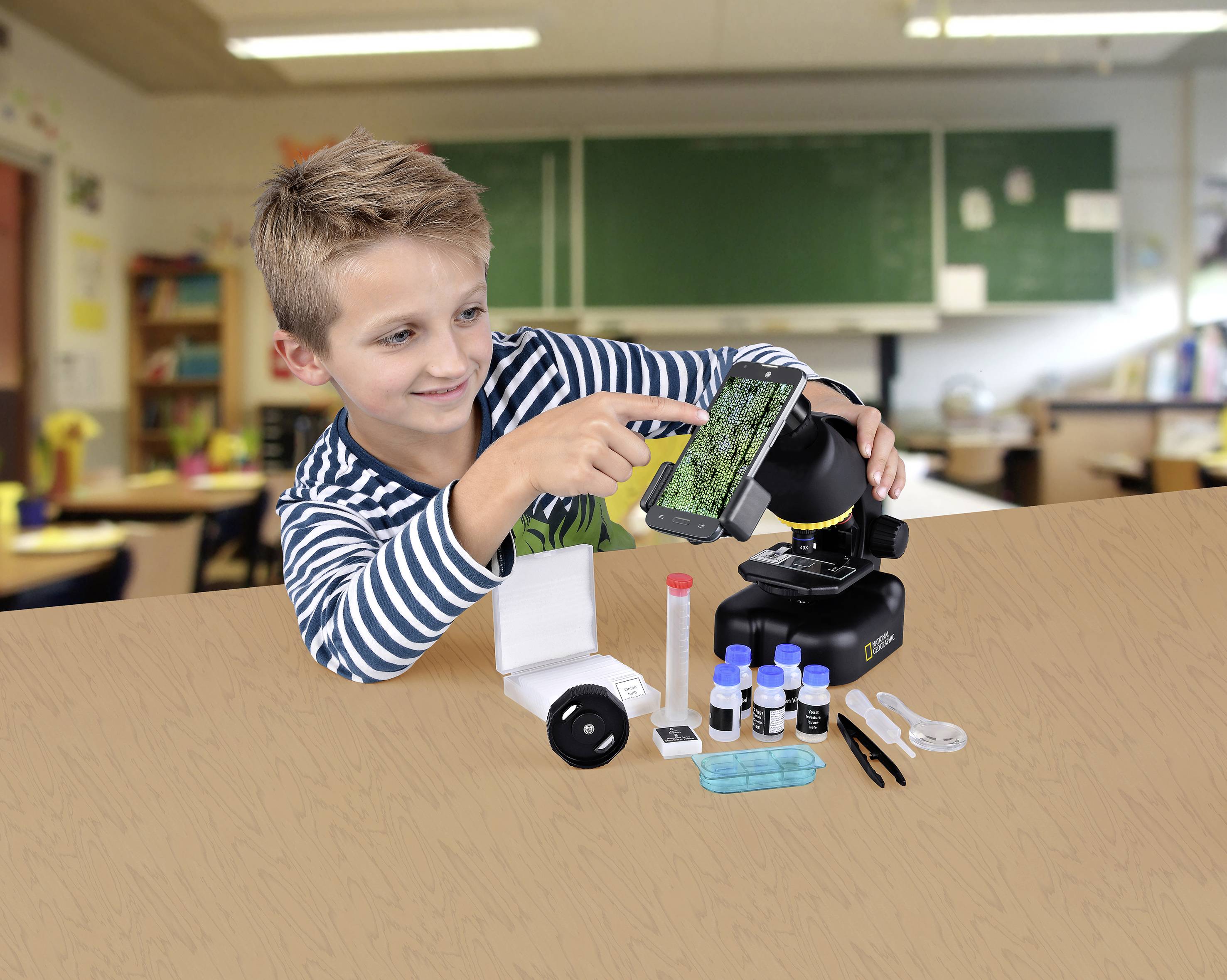 Chłopiec korzysta z zestawu mikroskopowego z akcesoriami w sali lekcyjnej. Zainteresowanym wzrokiem patrzy na smartfon podłączony do mikroskopu.