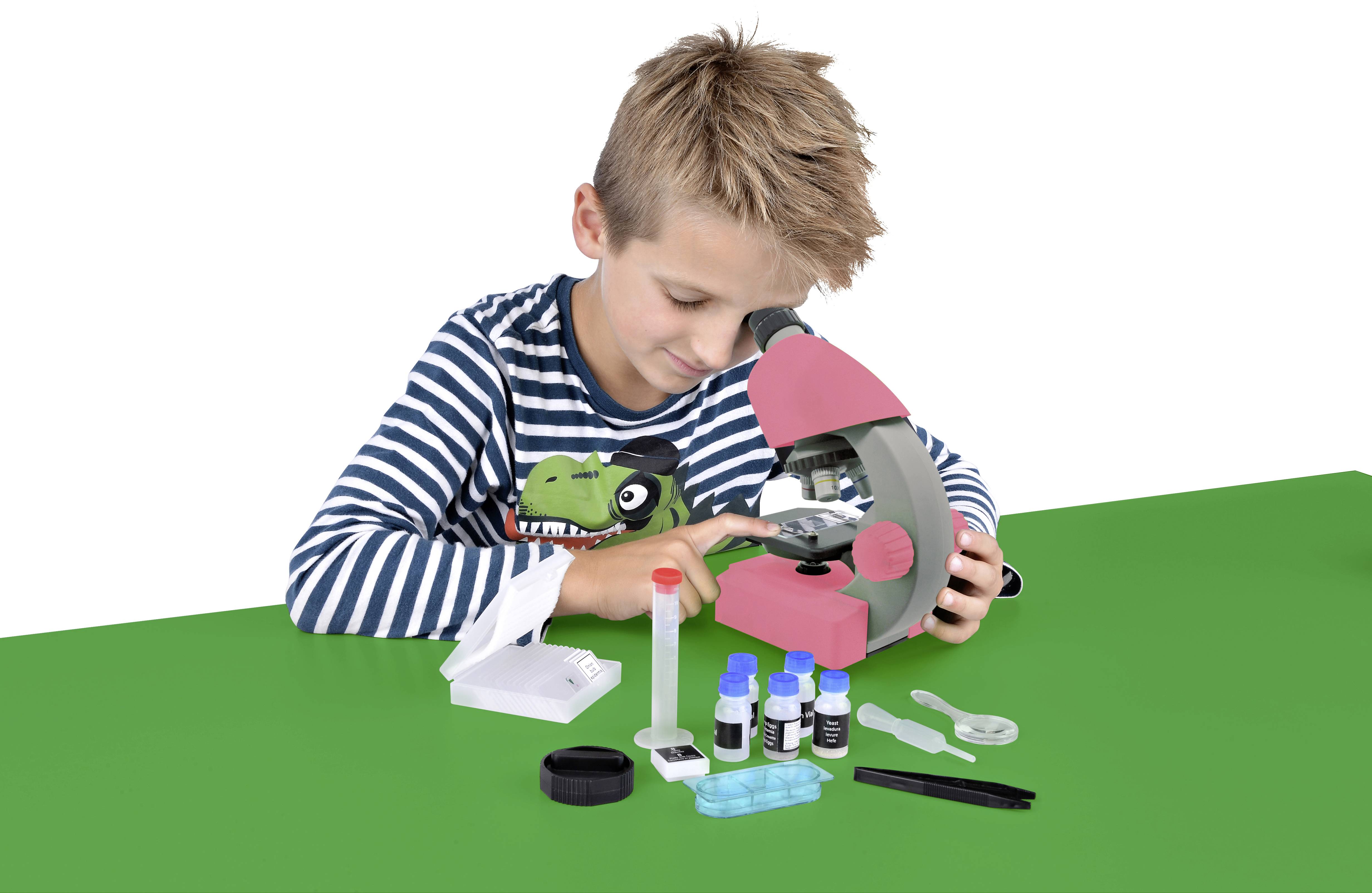 Chłopiec przygląda się obiektowi przez mikroskop ustawiony na zielonym stole. Przed nim rozłożony jest sprzęt laboratoryjny i próbki.