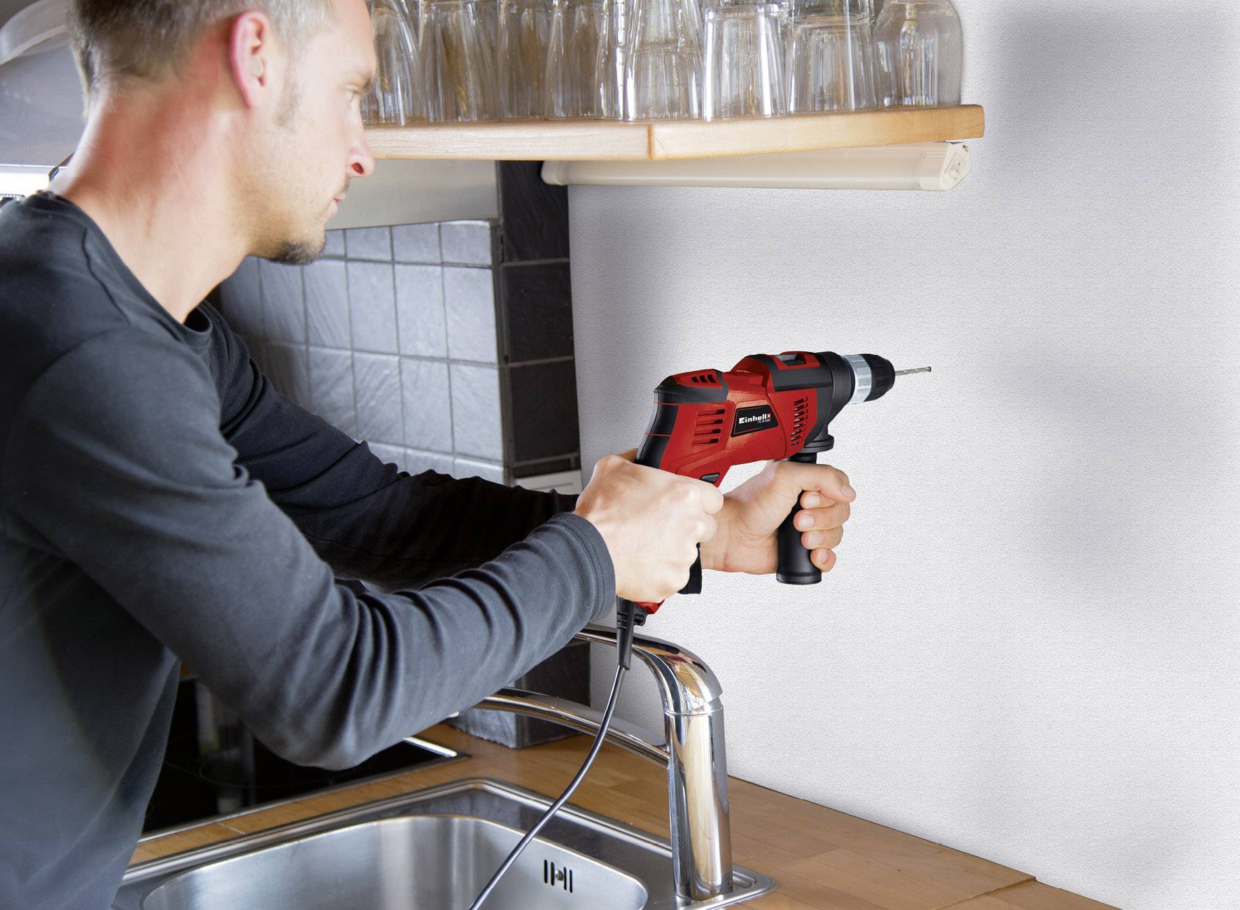 Mężczyzna wierca dziurę w ścianie nad zlewem kuchennym za pomocą czerwonej wiertarki.