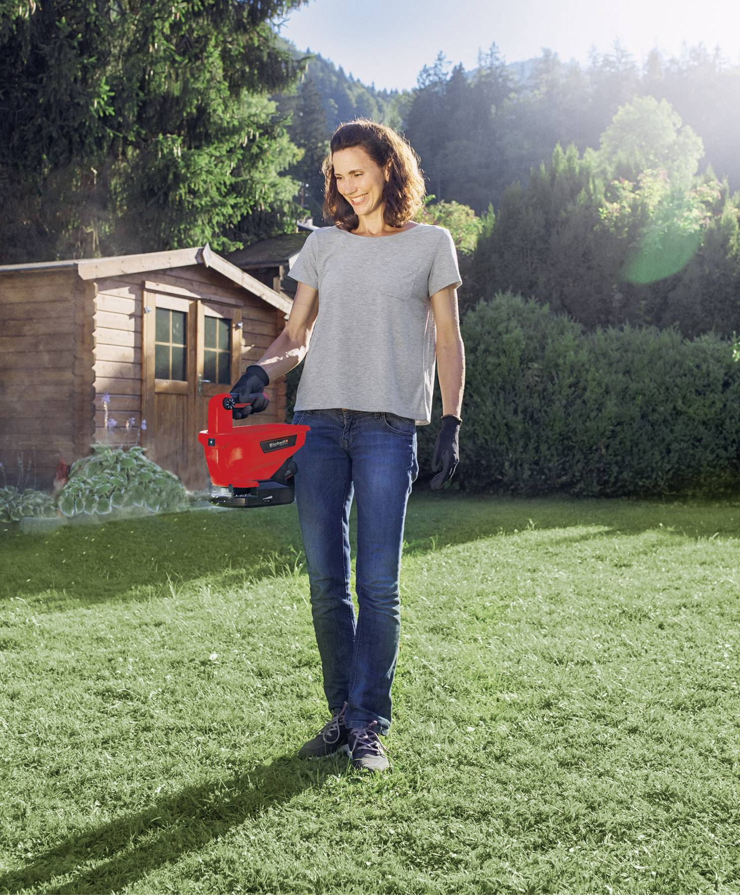 Kobieta stoi w ogrodzie, trzymając czerwoną kosiarką, otoczona zielonymi drzewami i drewnianym składzikiem w tle.