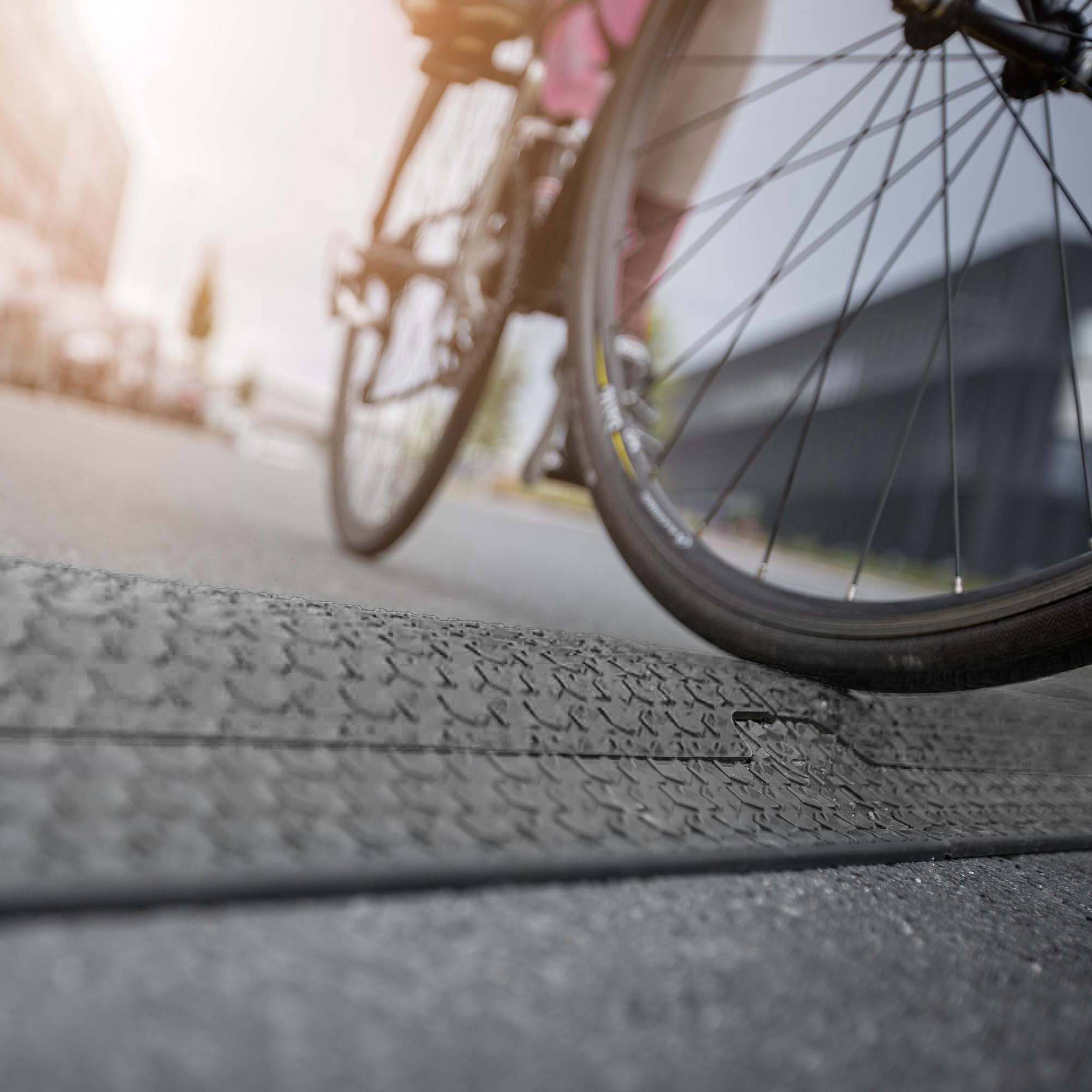 Rowerzysta przejeżdża przez krawężnikowy podjazd, który nachylony prowadzi na ulicę. W tle widać nieostre budynki.
