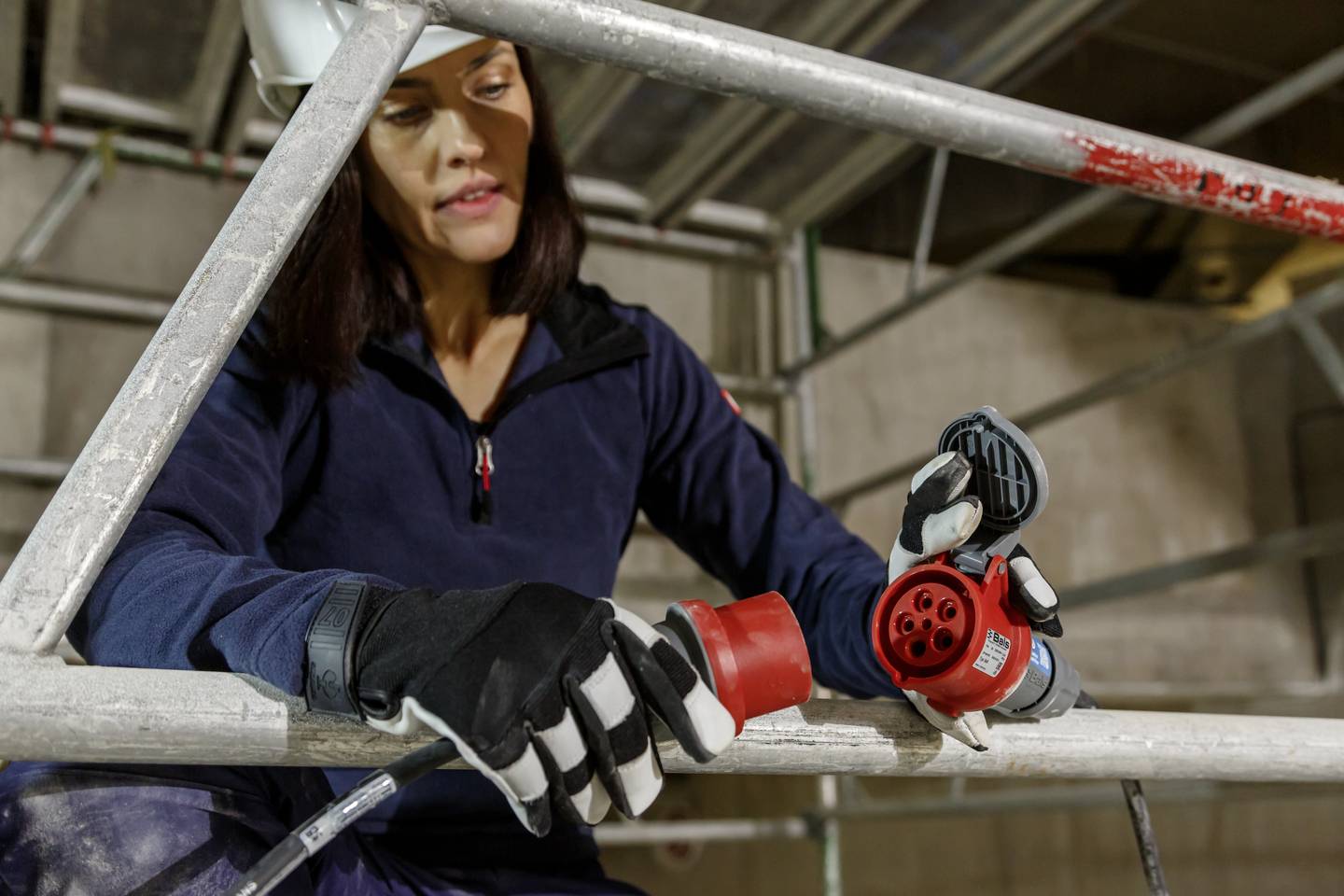 Kobieta w stroju roboczym i kasku łączy czerwony kabel elektryczny na budowie. Ma na sobie czarne rękawice.