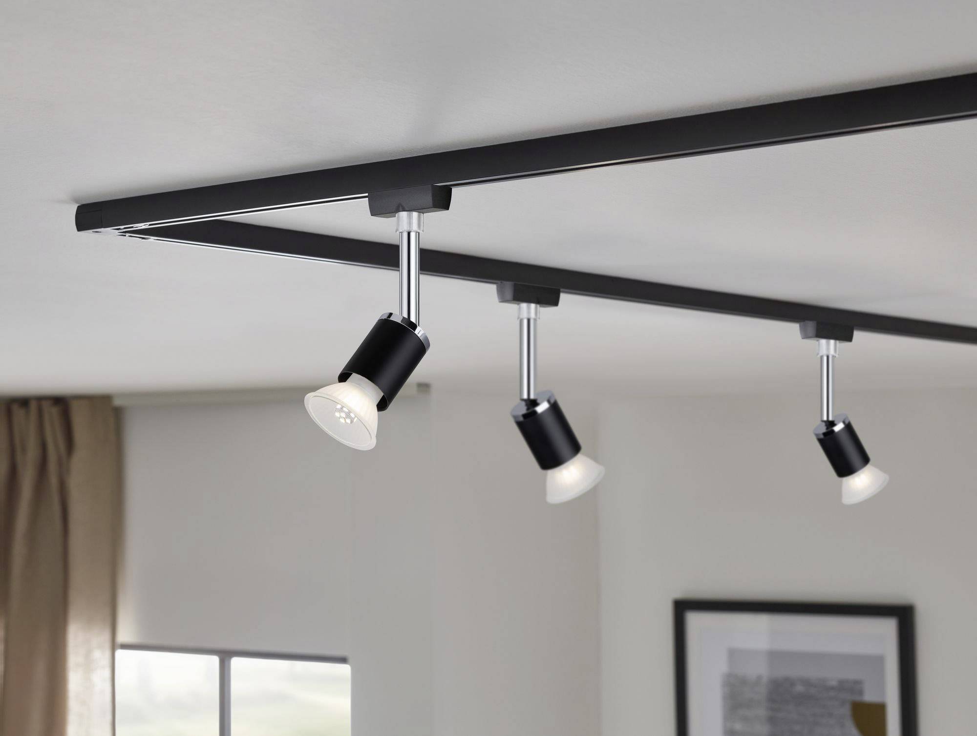 Trzy reflektory sufitowe zamontowane na szynie oświetlają pomieszczenie. W tle widoczne jest okno z zasłoną oraz obraz w ramie.