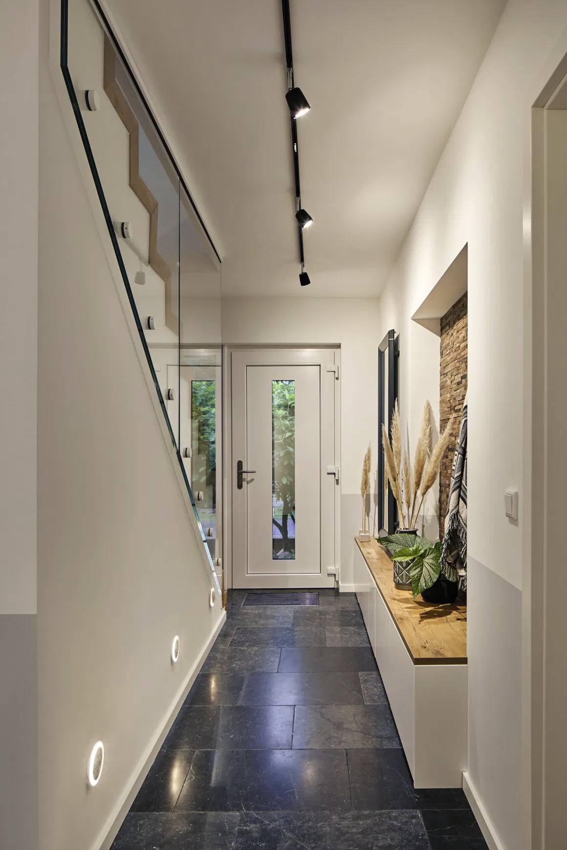 Nowoczesne wnętrze z drewnianą podłogą, zieloną ścianą pokrytą roślinnością oraz szafą z drewnianymi drzwiami. Okno dachowe zapewnia dużo naturalnego światła.