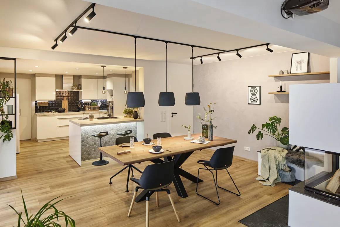 Nowoczesny salon z drewnianym stołem jadalnianym, czarnymi krzesłami i wiszącymi lampami. W tle kanapa, rośliny i telewizor na ścianie.