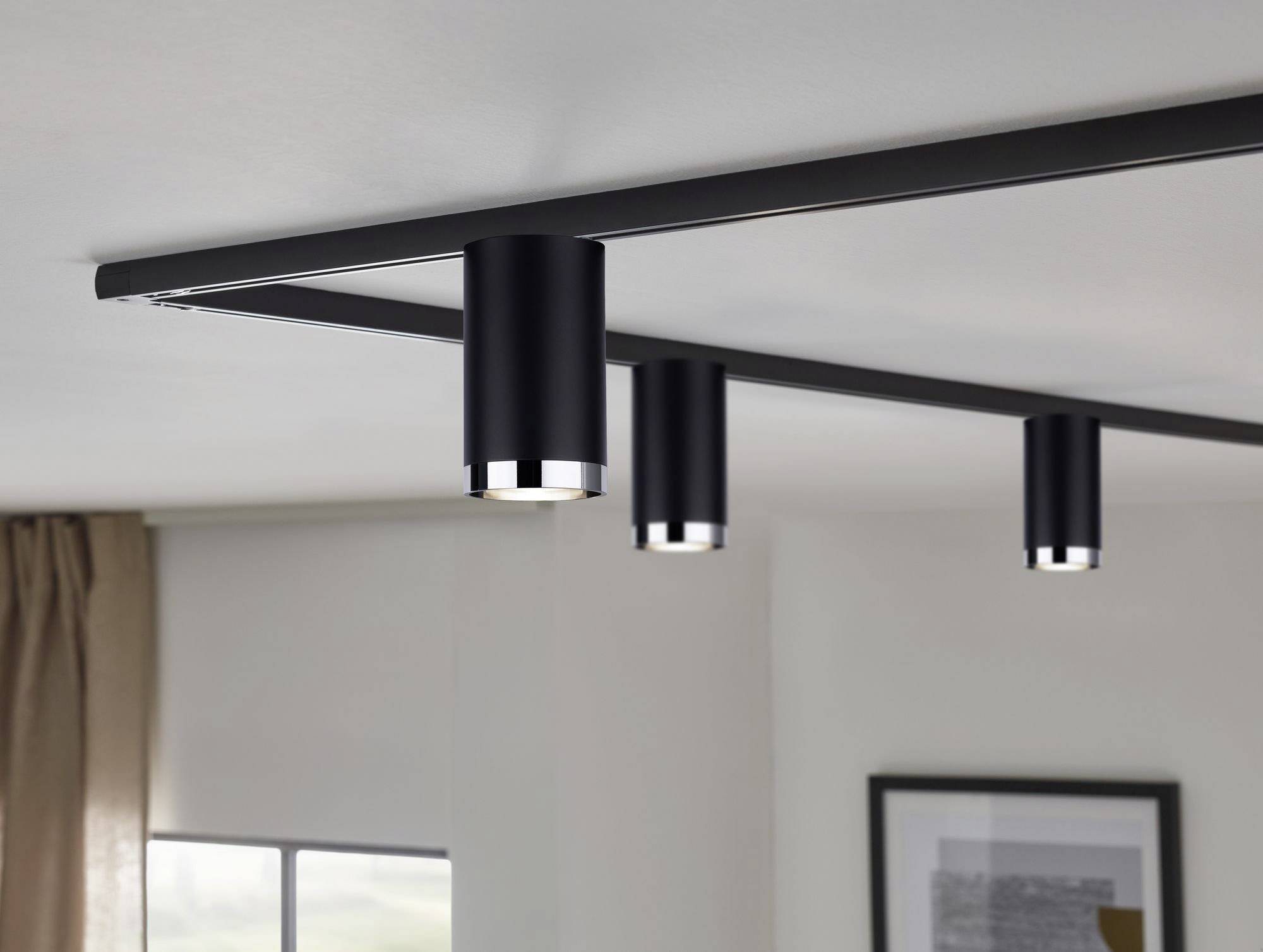 Czarne, cylindryczne lampy sufitowe zamontowane na szynie w nowoczesnym pomieszczeniu; oświetlają przestrzeń pod sobą.