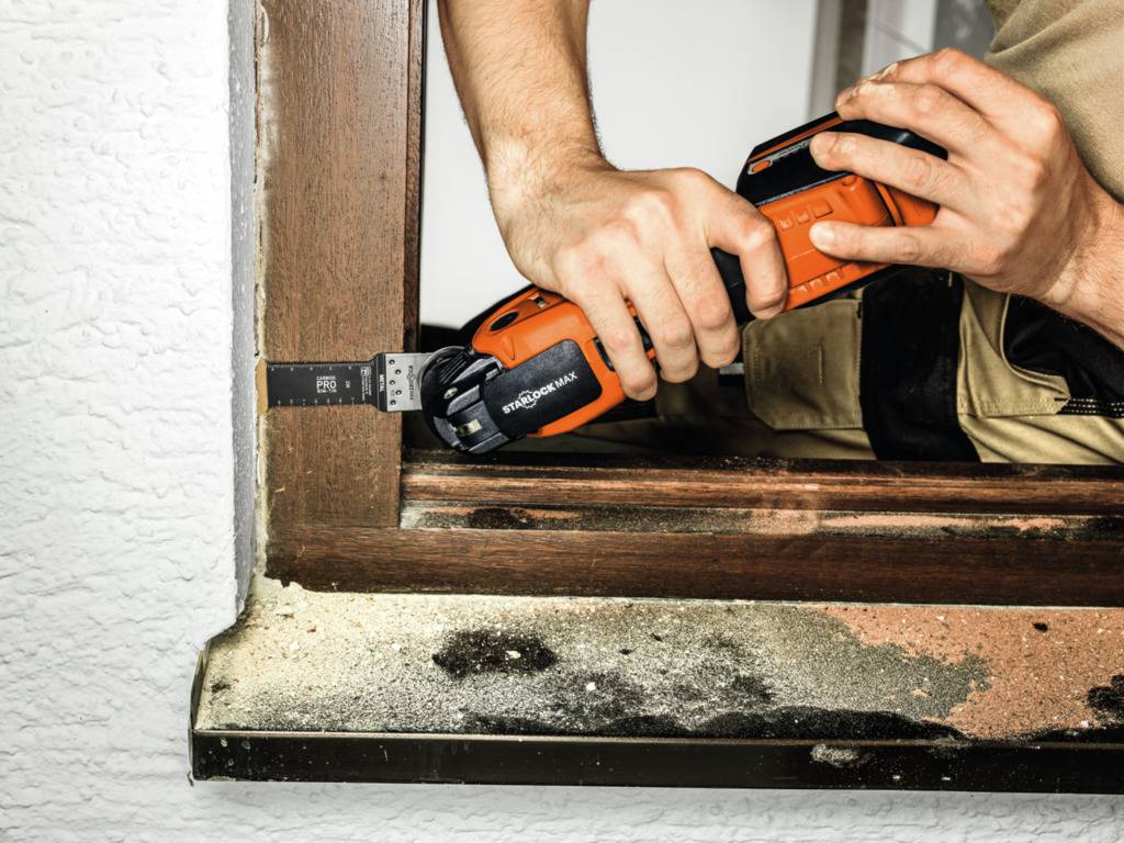 Osoba używa narzędzia oscylacyjnego do obróbki otworu drewnianej ramy okiennej. Widoczny jest pył i wióry.