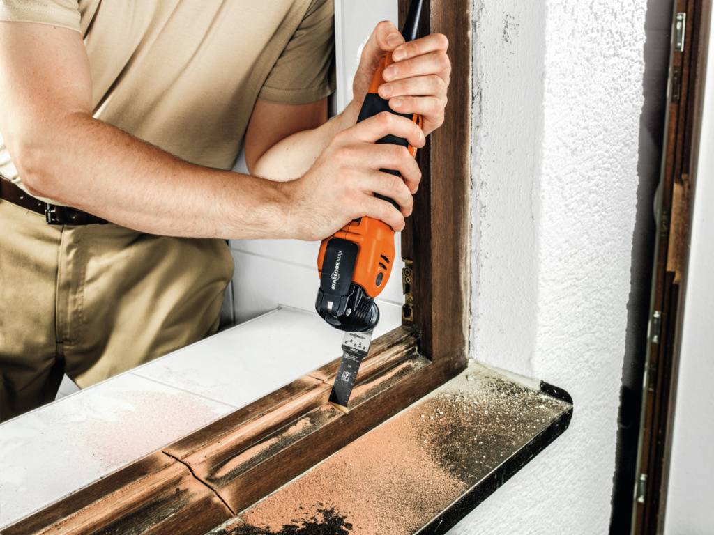 Osoba używa narzędzia oscylacyjnego do naprawy okna. Wióry drewniane leżą na parapecie.