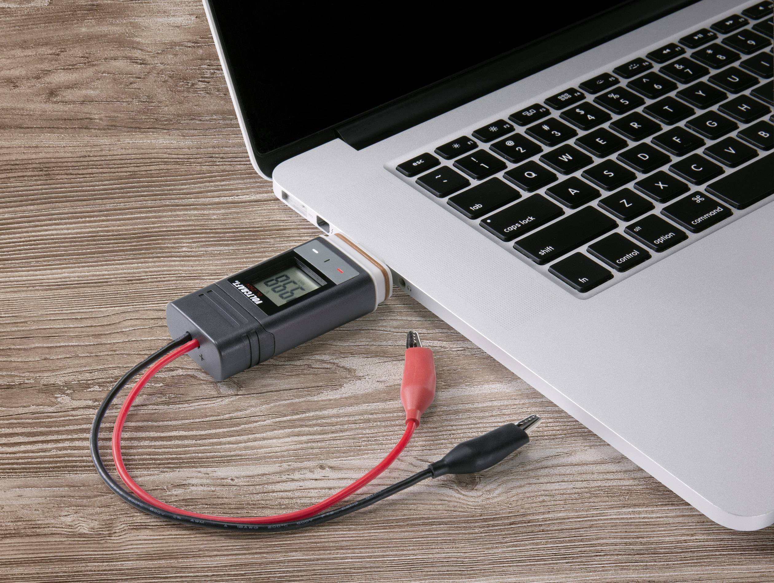 Laptop na drewnianym stole z podłączonym miernikiem napięcia, sprawdzającym napięcie poprzez port USB.