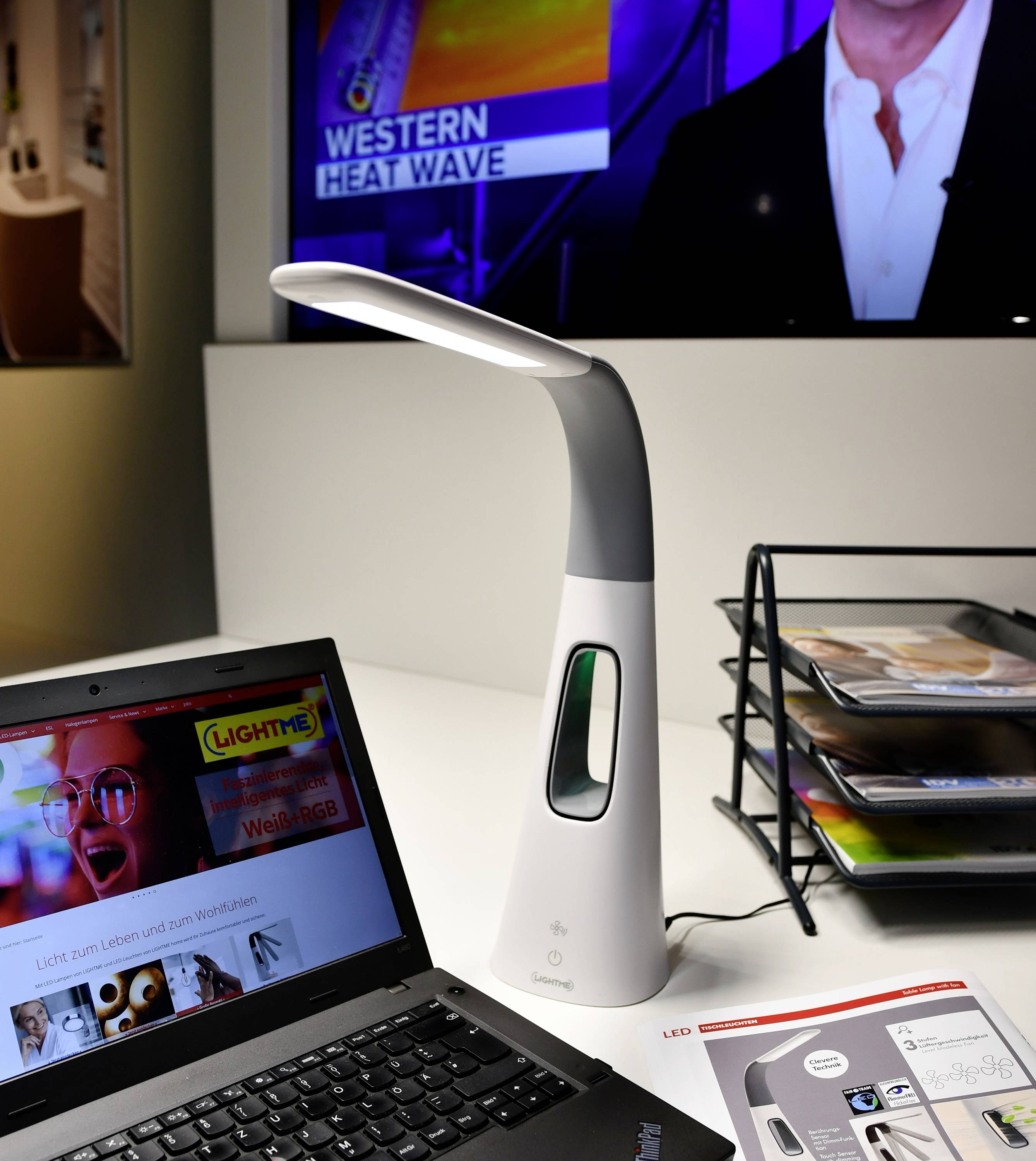 Stanowisko pracy technicznego specjalisty z laptopem, nowoczesną lampką biurkową oraz telewizorem w tle, na którym wyświetlana jest audycja informacyjna.