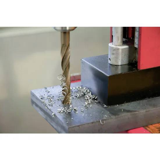 Wiertarka stacjonarna aktywnie wierci otwór w metalowej płycie, a wokół wiertła rozrzucone są metalowe wióry, świadczące o niedawnym użyciu.