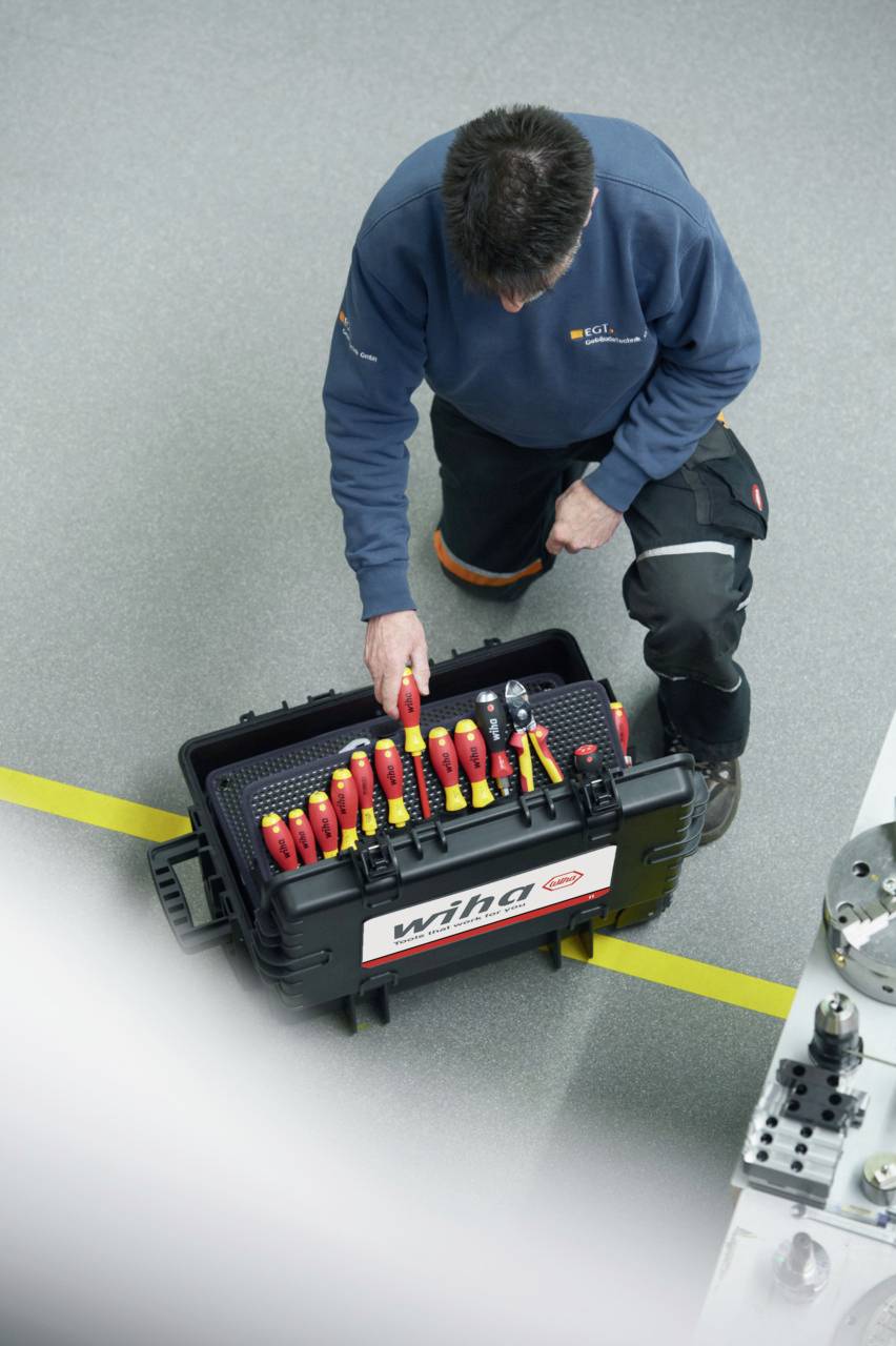 Technik klęczy obok otwartej walizki narzędziowej na szarej podłodze. Walizka zawiera różne narzędzia.