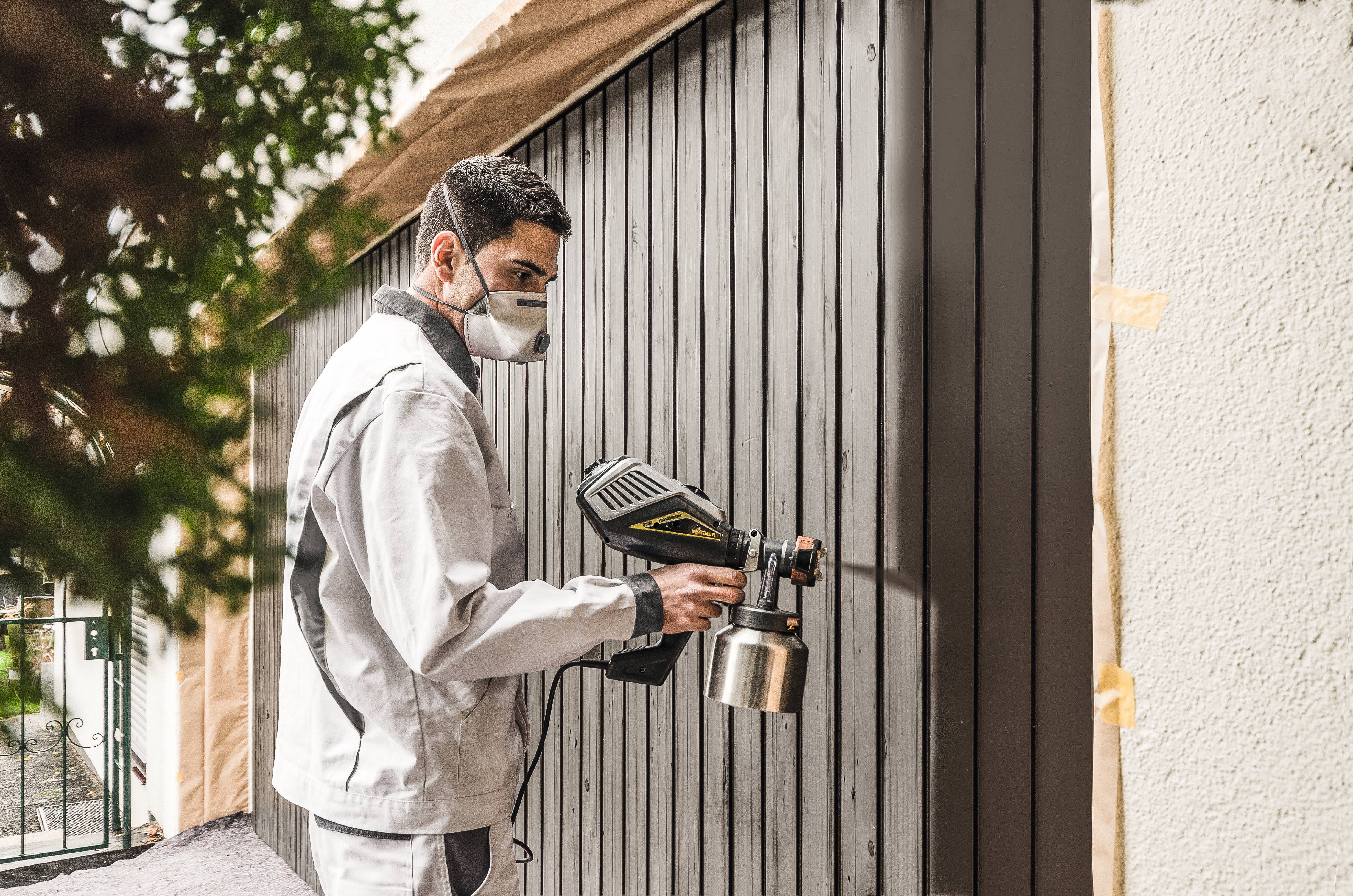 Mężczyzna w odzieży roboczej maluje farbą bramę garażową. Nosi maskę ochronną i używa pistoletu natryskowego.