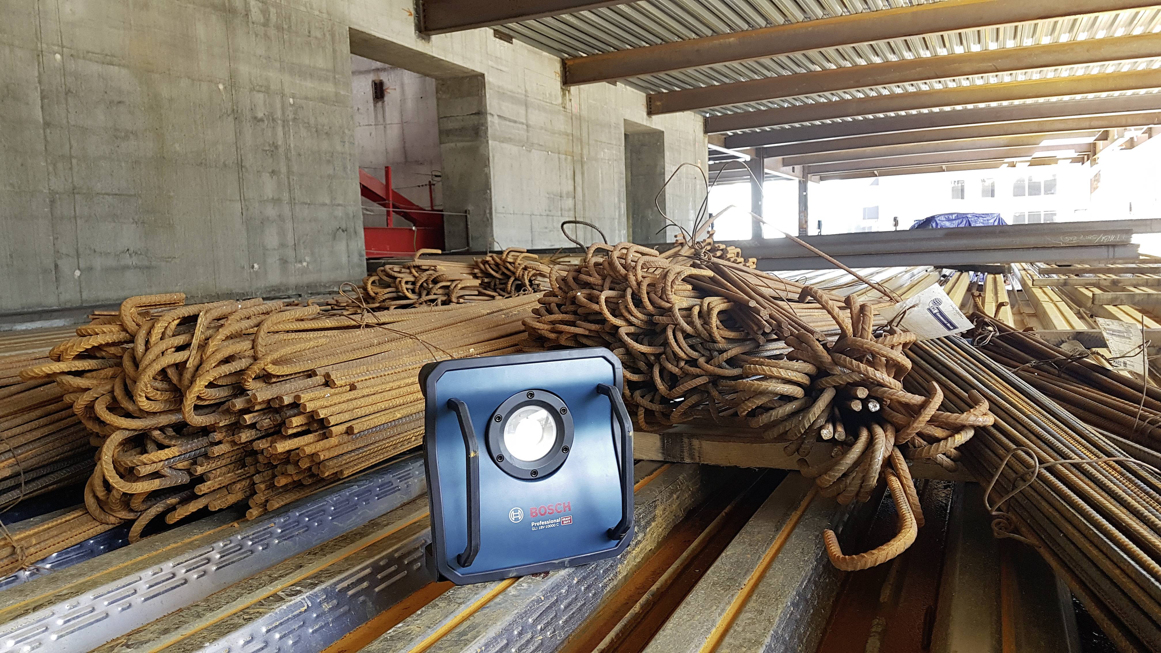 Zardzewiałe pręty stalowe są ułożone w stosach w hali magazynowej, a niebieski reflektor budowlany znajduje się na belce w pierwszym planie.