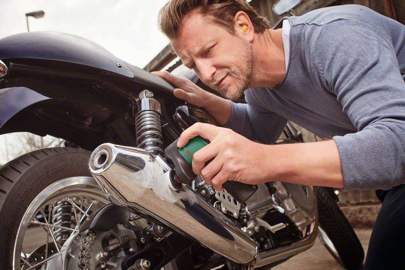 Mężczyzna kontroluje tylne koło i tłumik motocykla. Wygląda na skoncentrowanego i trzyma w ręku narzędzie.